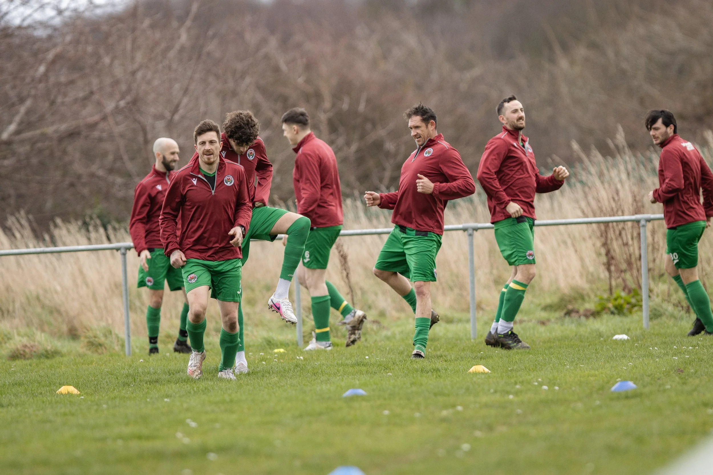 Group of male soccer players doing warm-up exercises on a grassy field, wearing red jackets and green shorts, with some players stretching and others jogging.