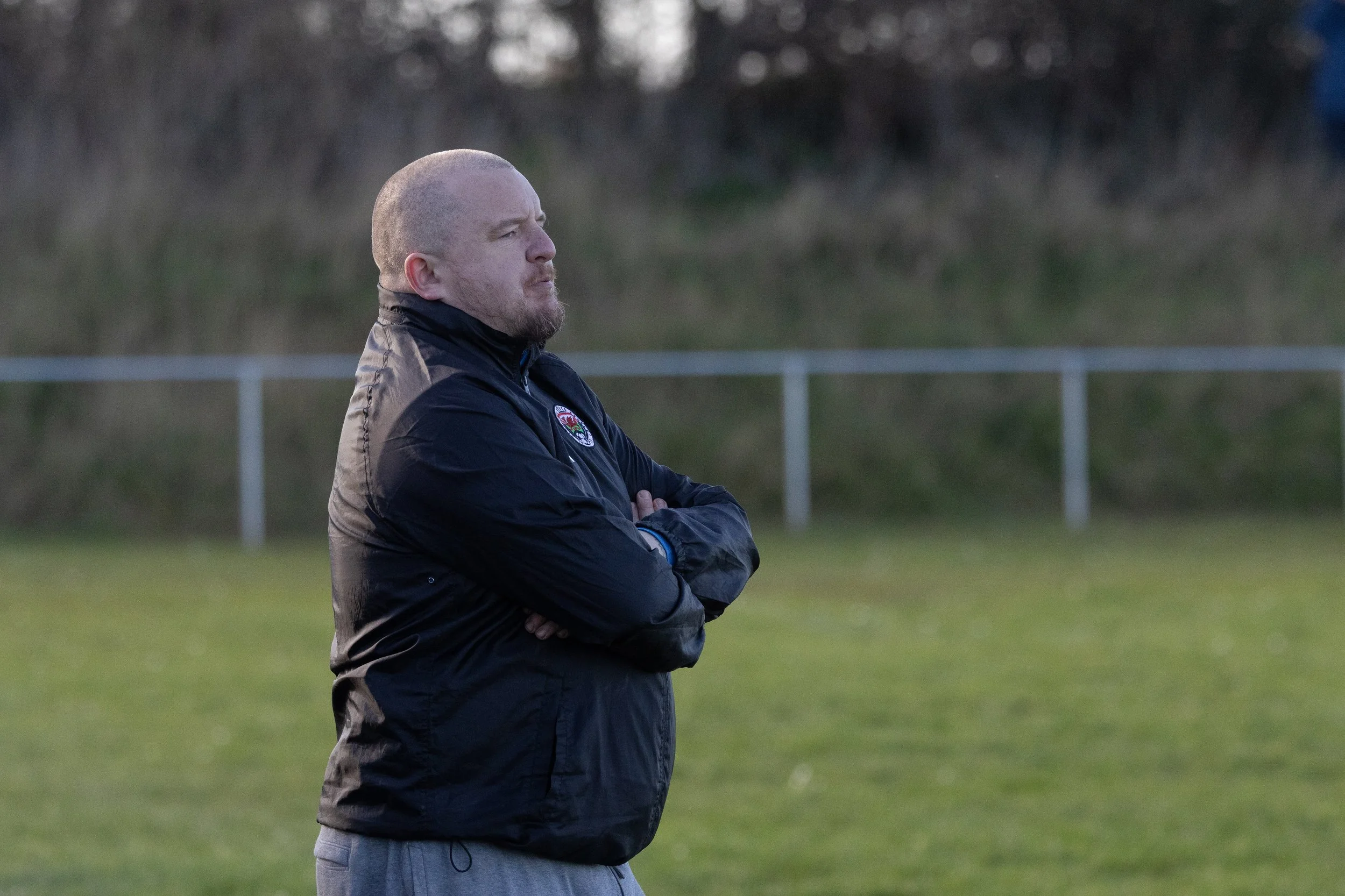 A man stands with arms crossed on a grassy field, wearing a black jacket, looking contemplative. The background shows a fence and blurred trees.