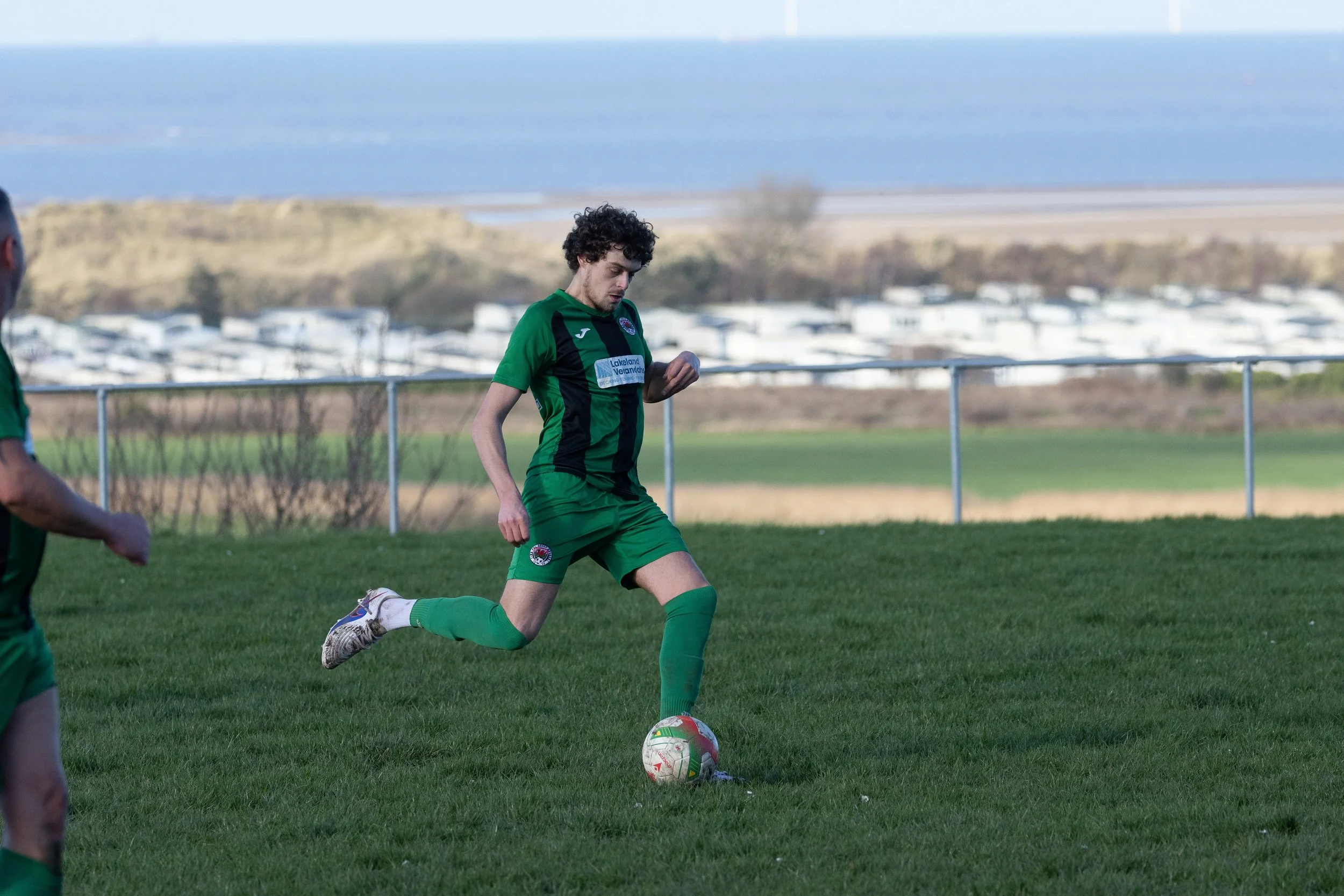A young male soccer player in a green and black uniform is about to kick a soccer ball on a grassy field. In the background, there are parked vehicles and a body of water.