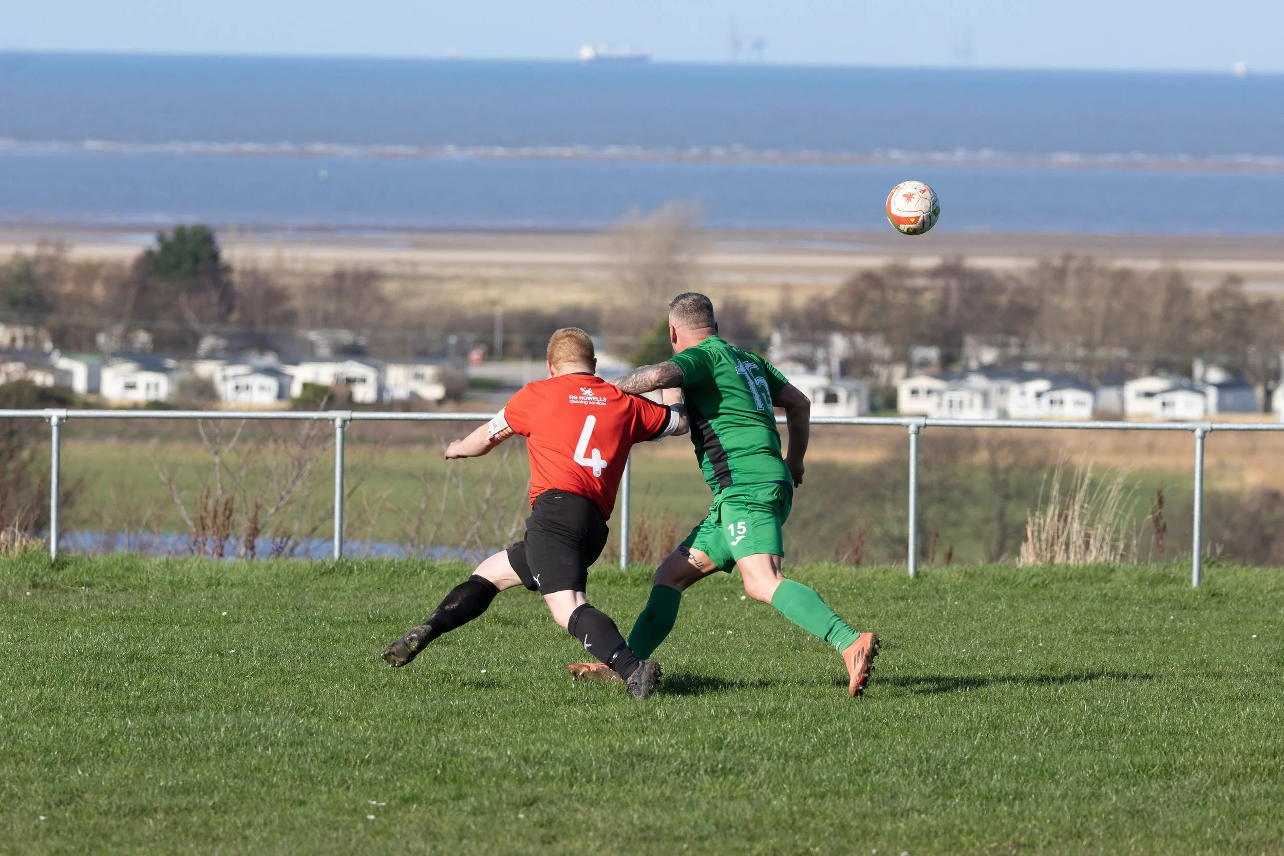 Two soccer players, one in a red jersey and another in a green jersey, compete for a ball on a grassy field with a view of the water and some distant buildings in the background.