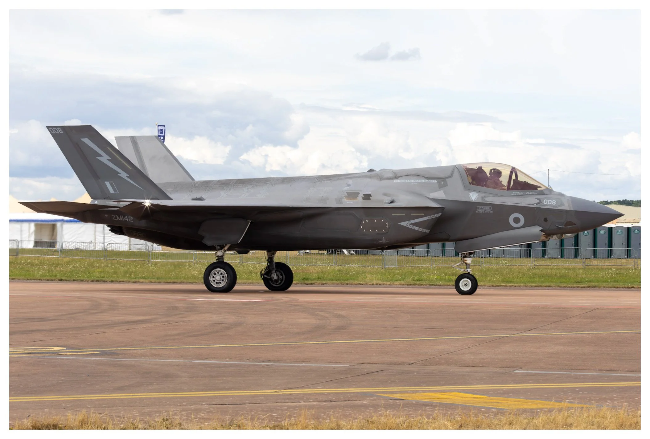 A modern fighter jet on runway with pilot visible in cockpit, grey camouflage pattern, under cloudy sky.