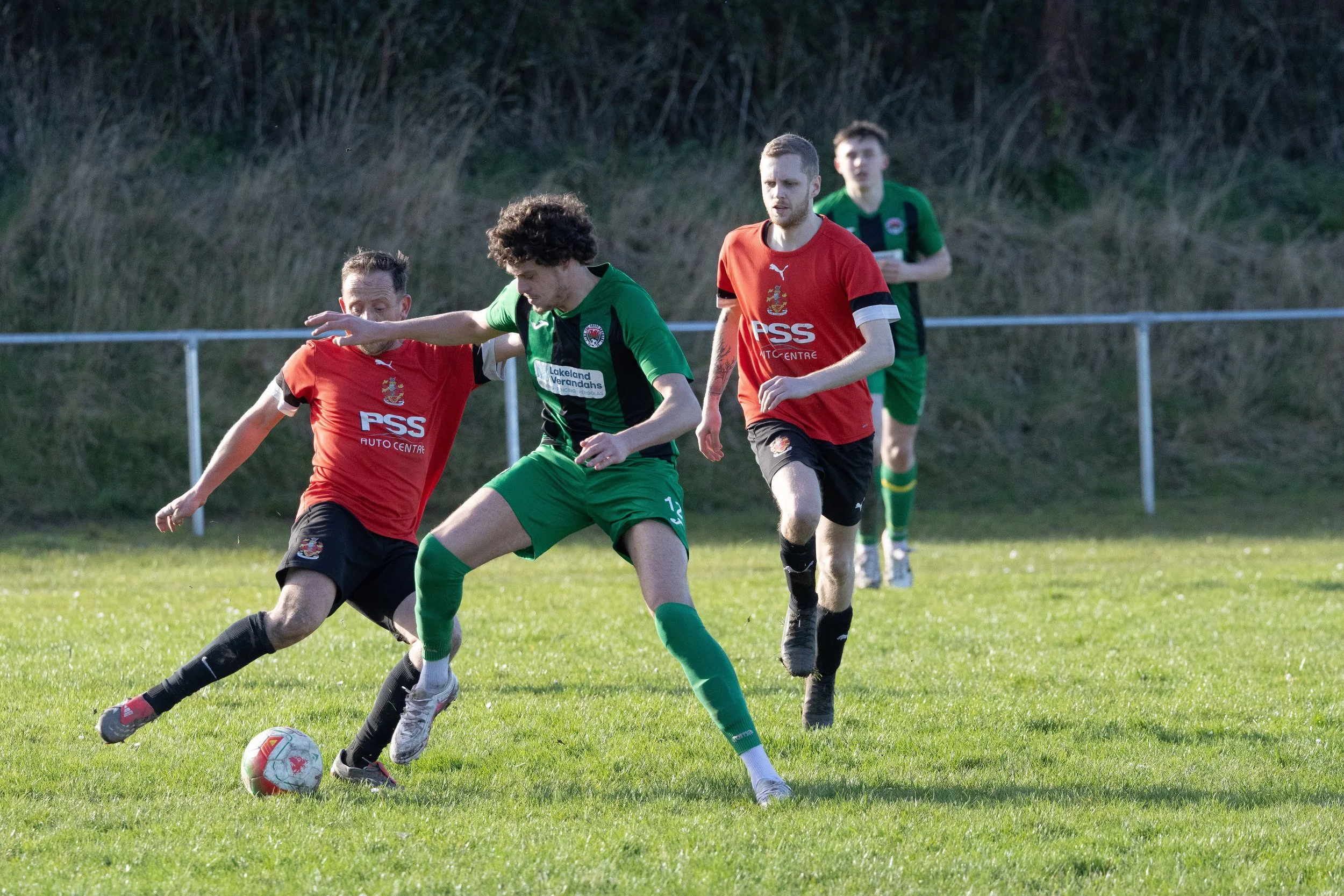 Four male soccer players during a match, two in red uniforms and two in green, competing for the ball on a grassy field with a hill and a fence in the background.