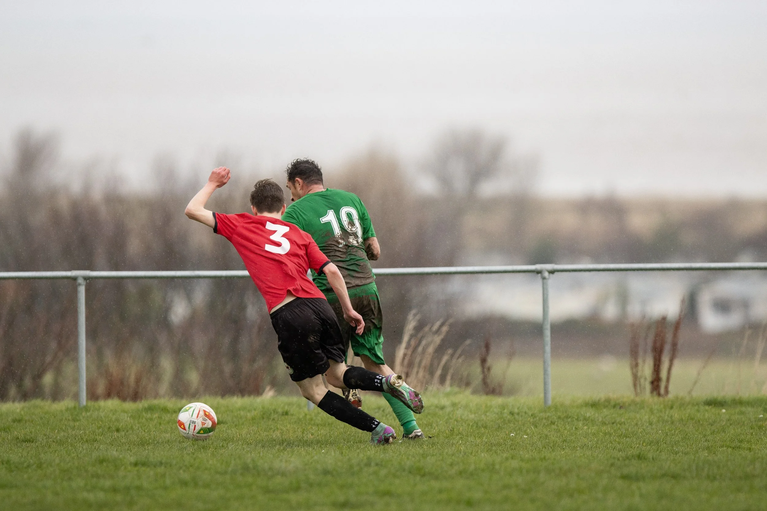 Two boys playing soccer on a grassy field in rainy weather, with one wearing a red shirt with number 3 and the other in a green shirt with number 19.