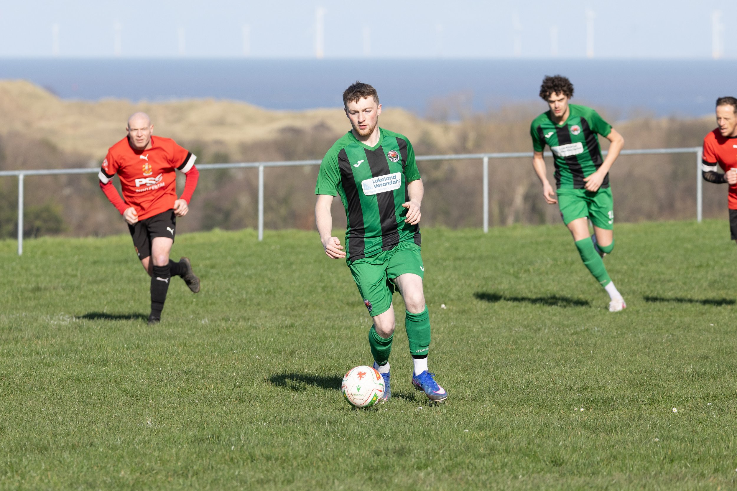 Soccer match with players in green and red jerseys on a grassy field outdoors, with wind turbines and hills visible in the background.