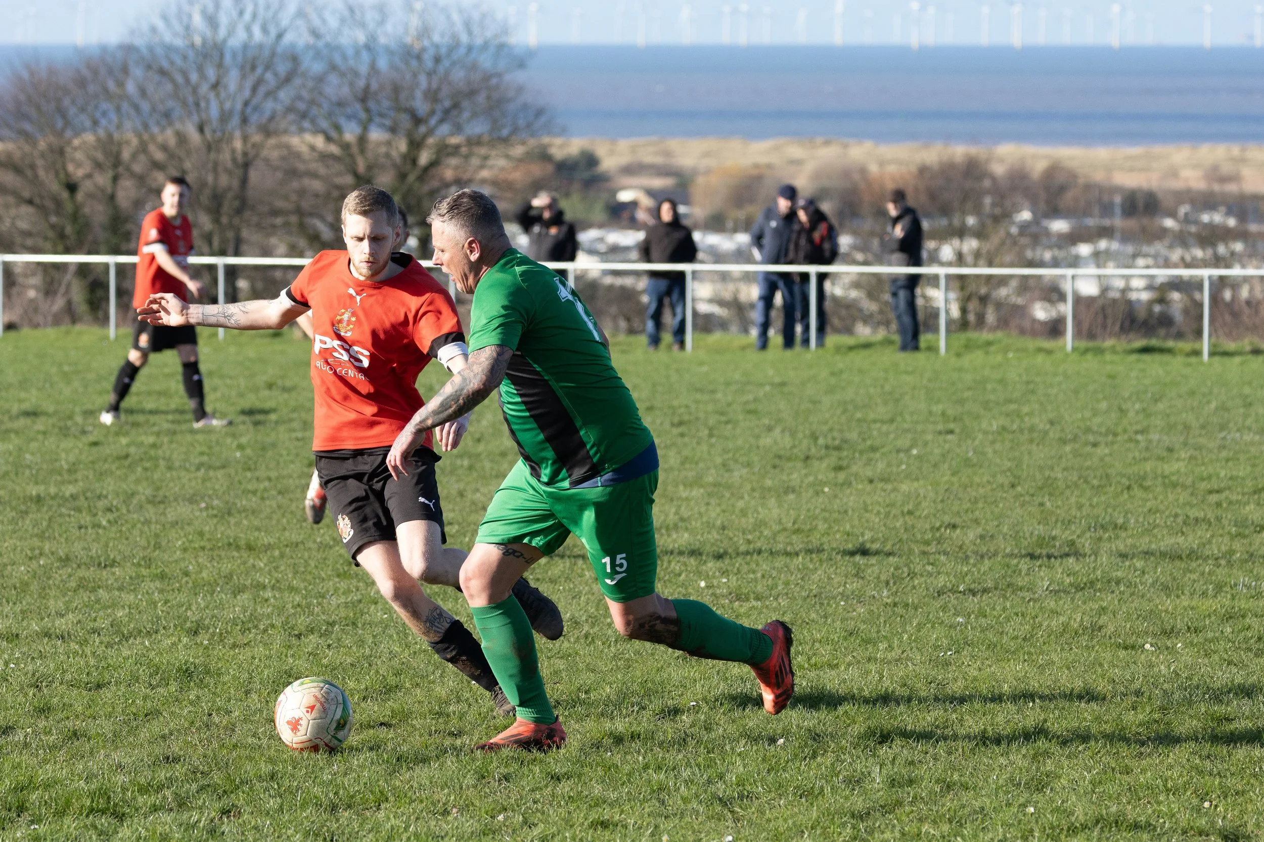 Two soccer players competing for the ball on a grassy field with a scenic background and spectators watching.