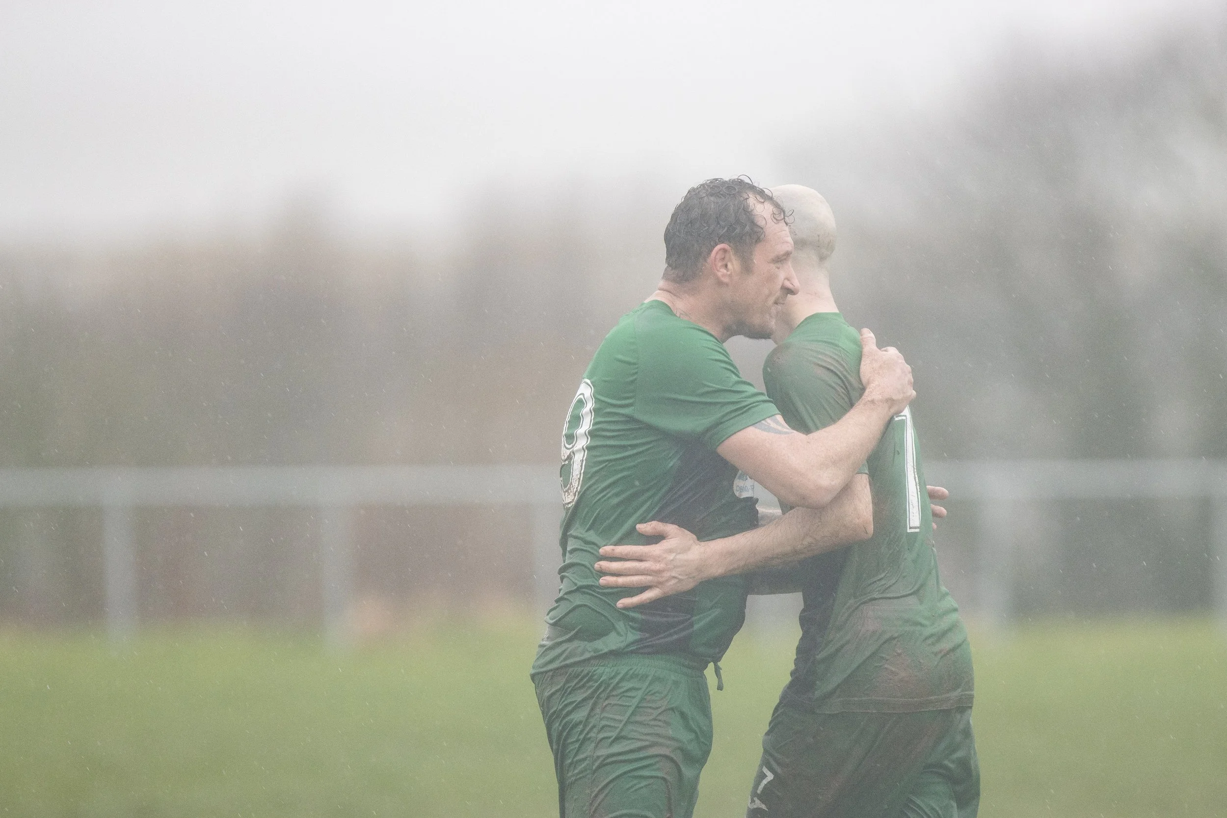 Two soccer players wearing green uniforms embrace in the rain on a grassy field.