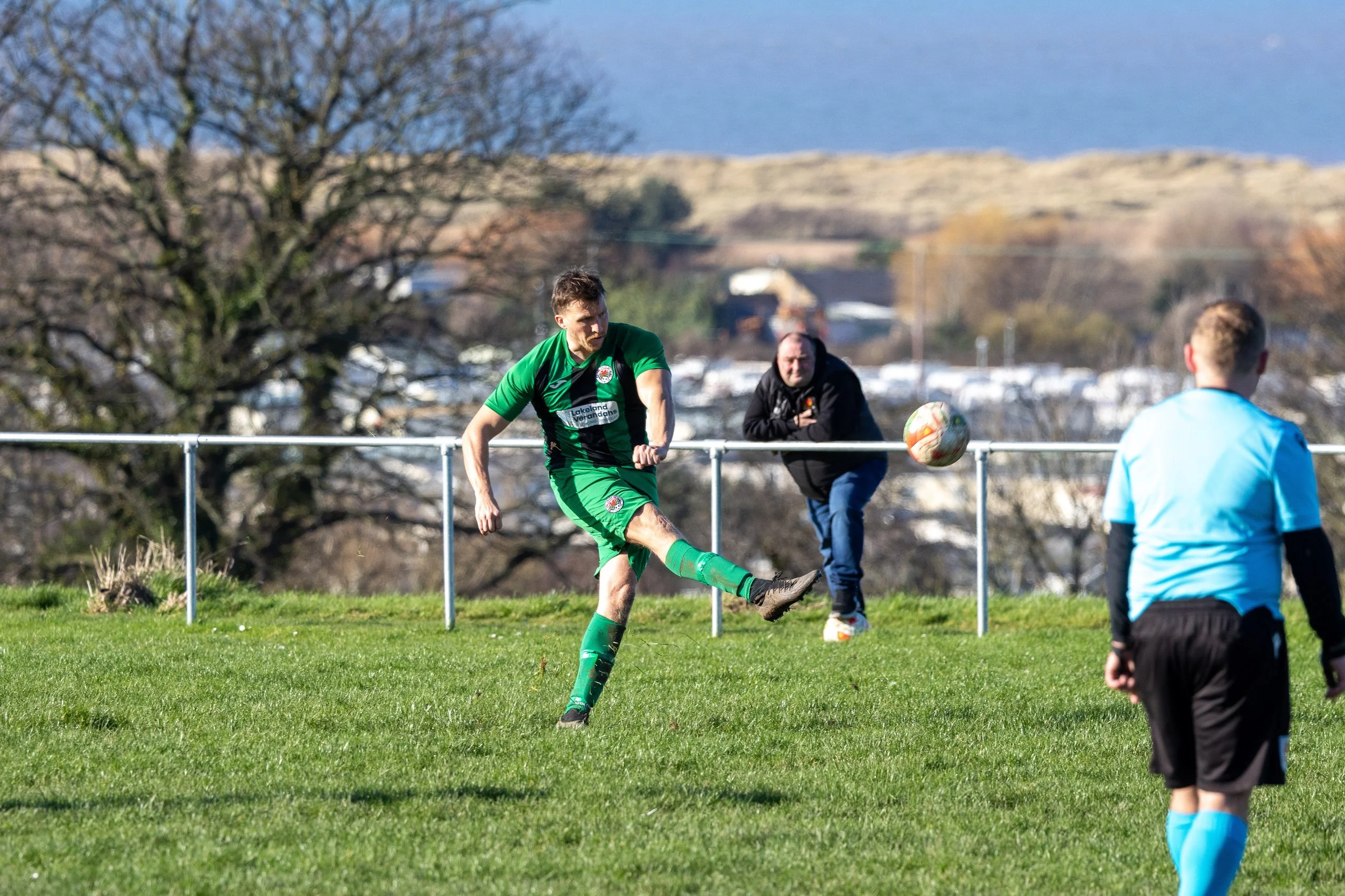 A soccer player in a green uniform kicking a ball during a match, with a referee in a light blue shirt and black shorts in the foreground and a spectator leaning on the fence watching in the background.
