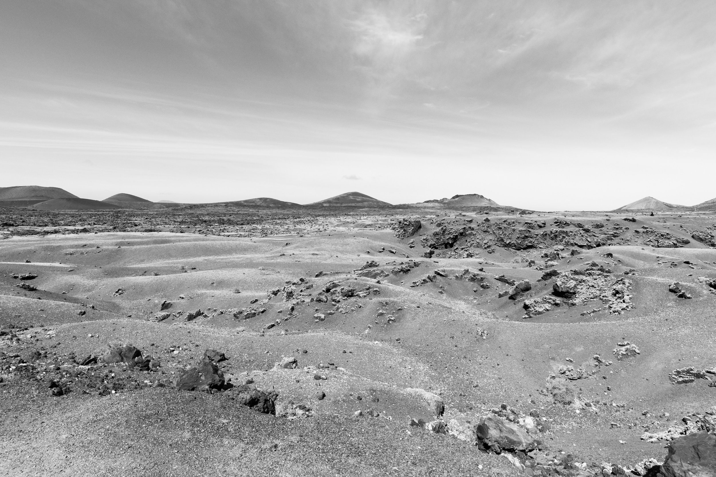 A barren, rocky desert landscape with small hills and mountains in the distance under a cloudy sky in black and white.