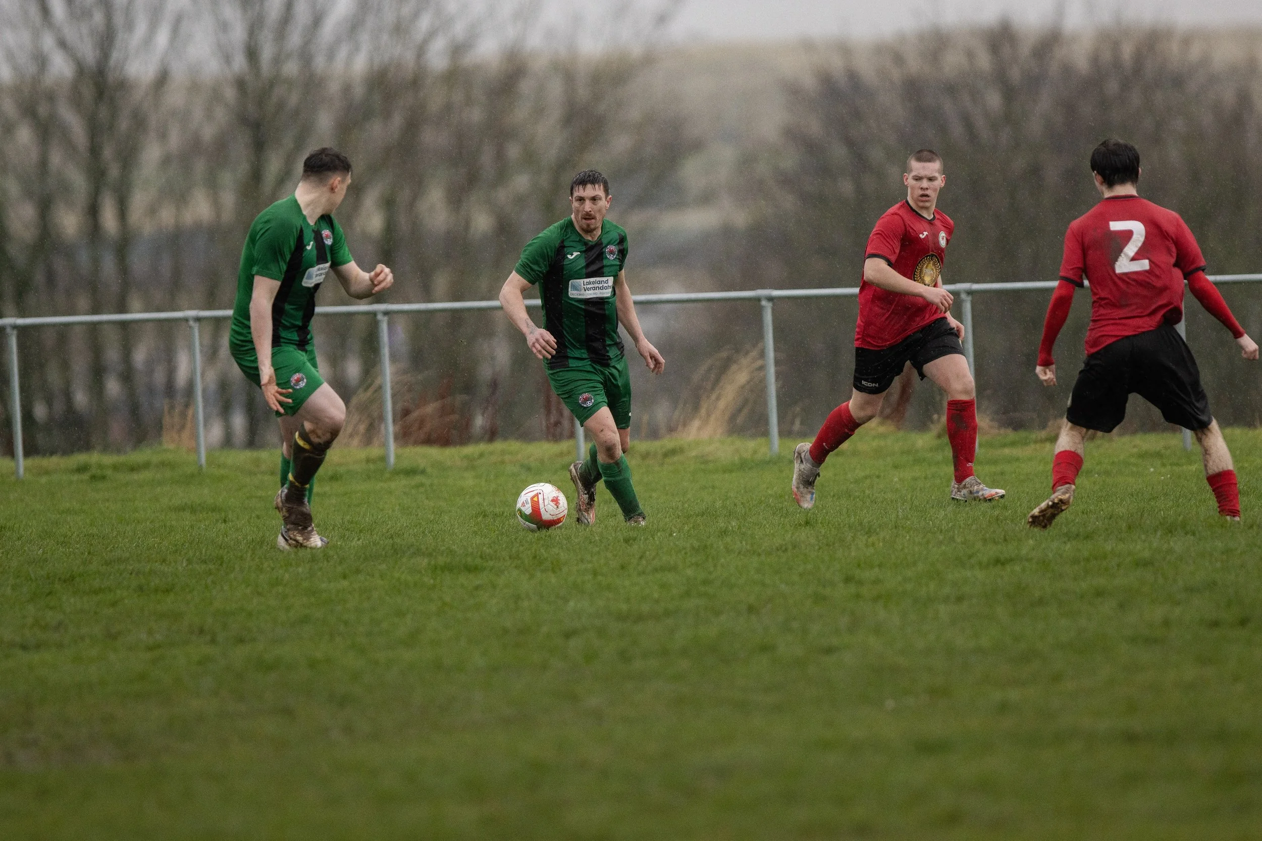 Four soccer players are on a grassy field during a match, with two wearing green uniforms and the other two in red and black uniforms, actively competing for the ball.