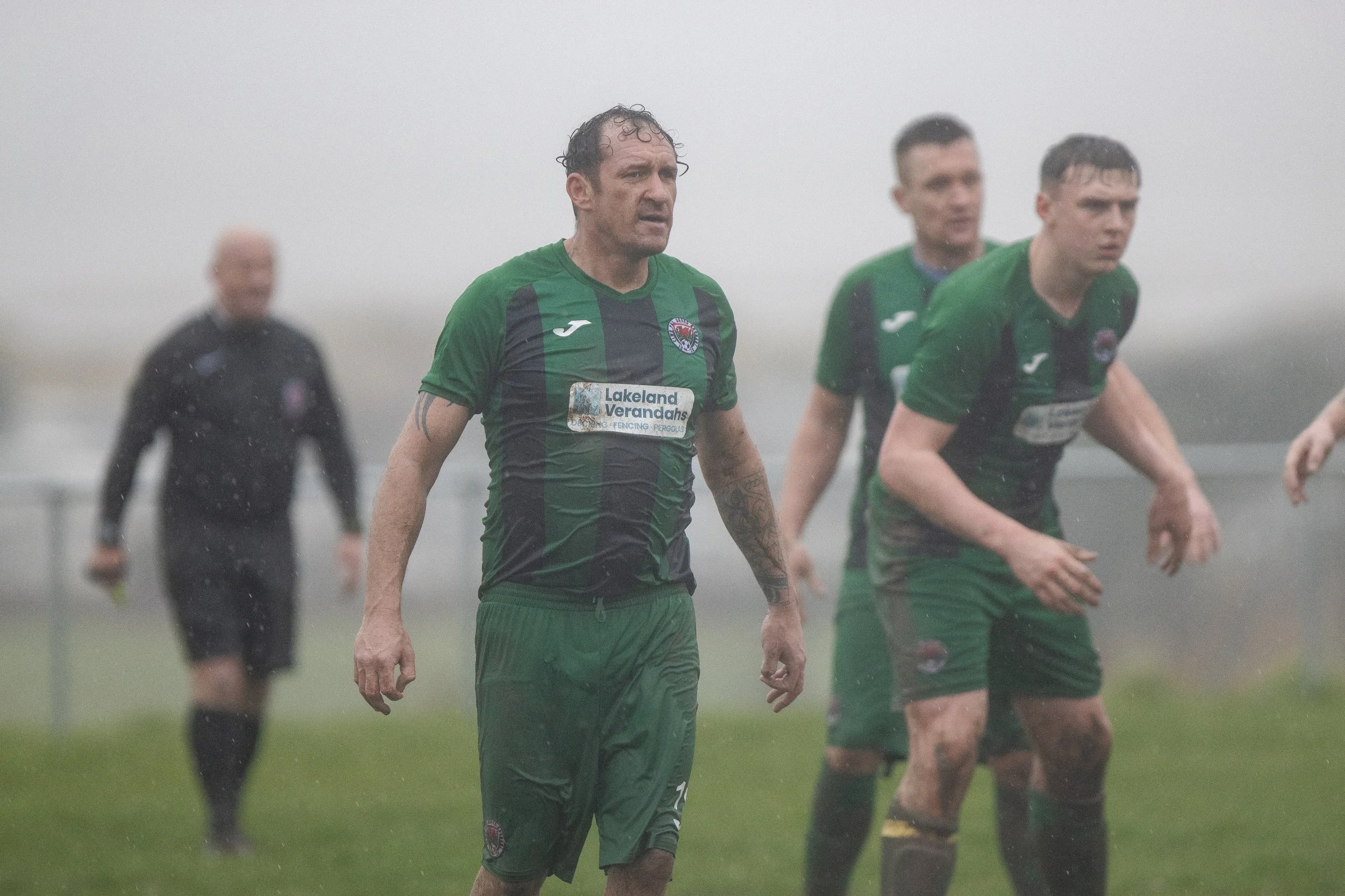 Soccer players and referee on a rainy, foggy field during a game, wearing green jerseys and dark shorts.