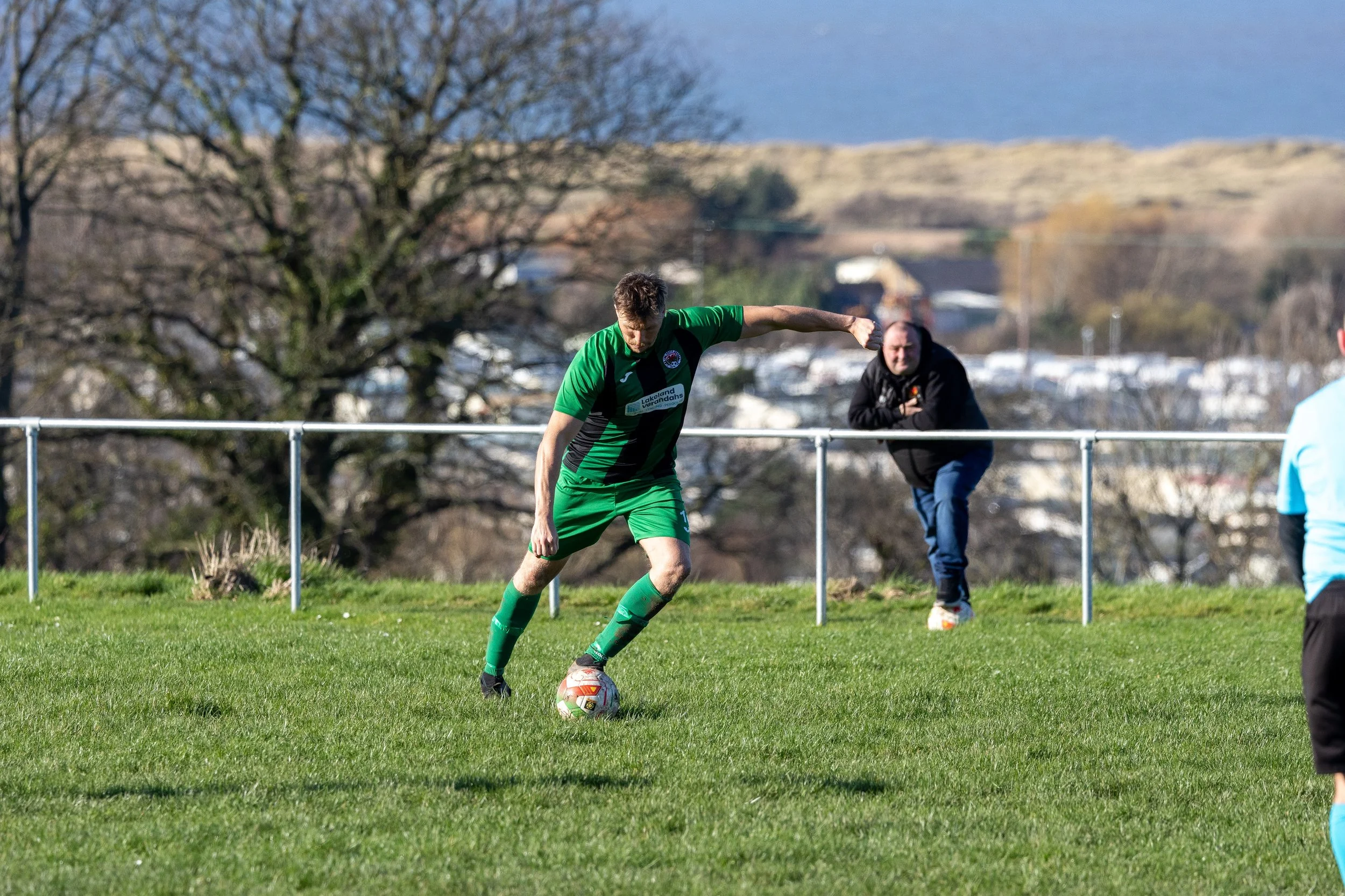 A soccer player in a green uniform about to kick a soccer ball on a grassy field, with a man watching behind a fence and trees in the background.