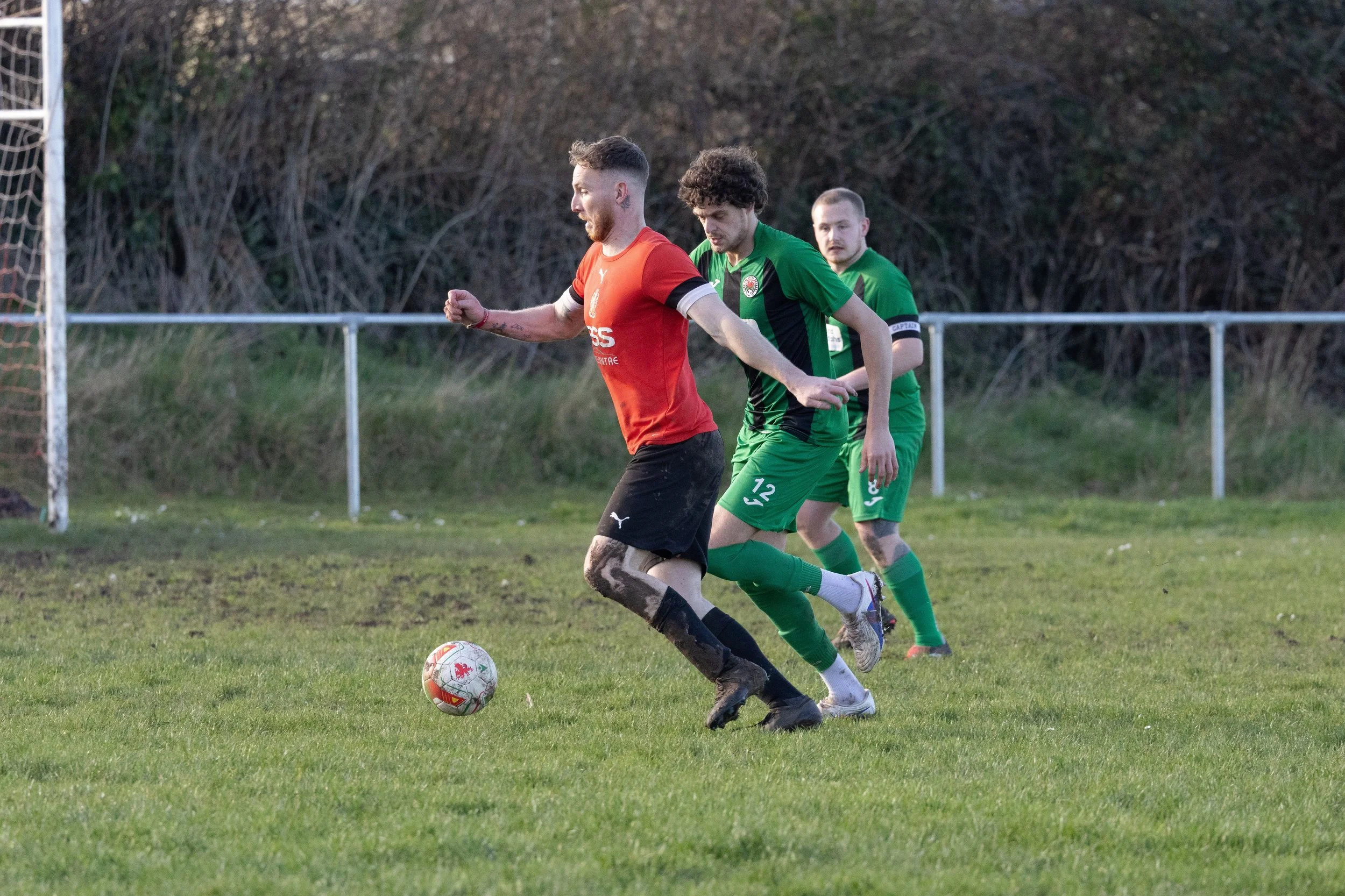 Three soccer players competing for the ball on a grassy field, with two in green jerseys and one in a red jersey, during daytime.