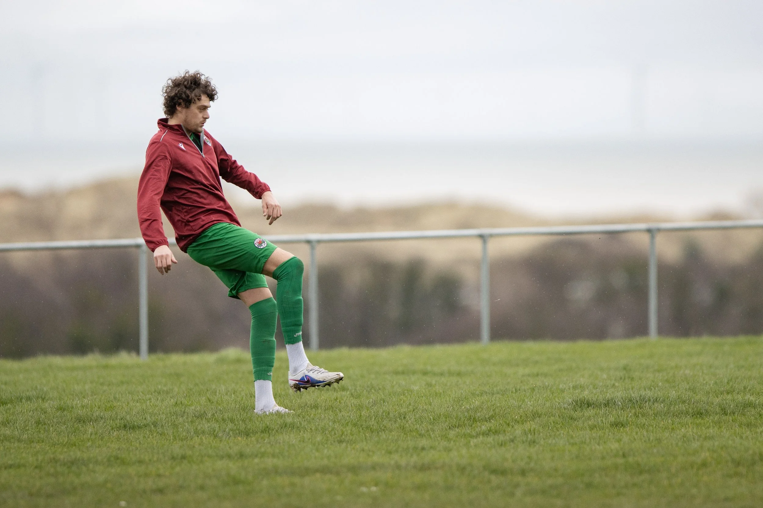A soccer player is on a grassy field in mid-motion, wearing a red jacket, green shorts, and green socks. He is lifting his right leg as if to kick or control a ball, with trees and a cloudy sky in the background.