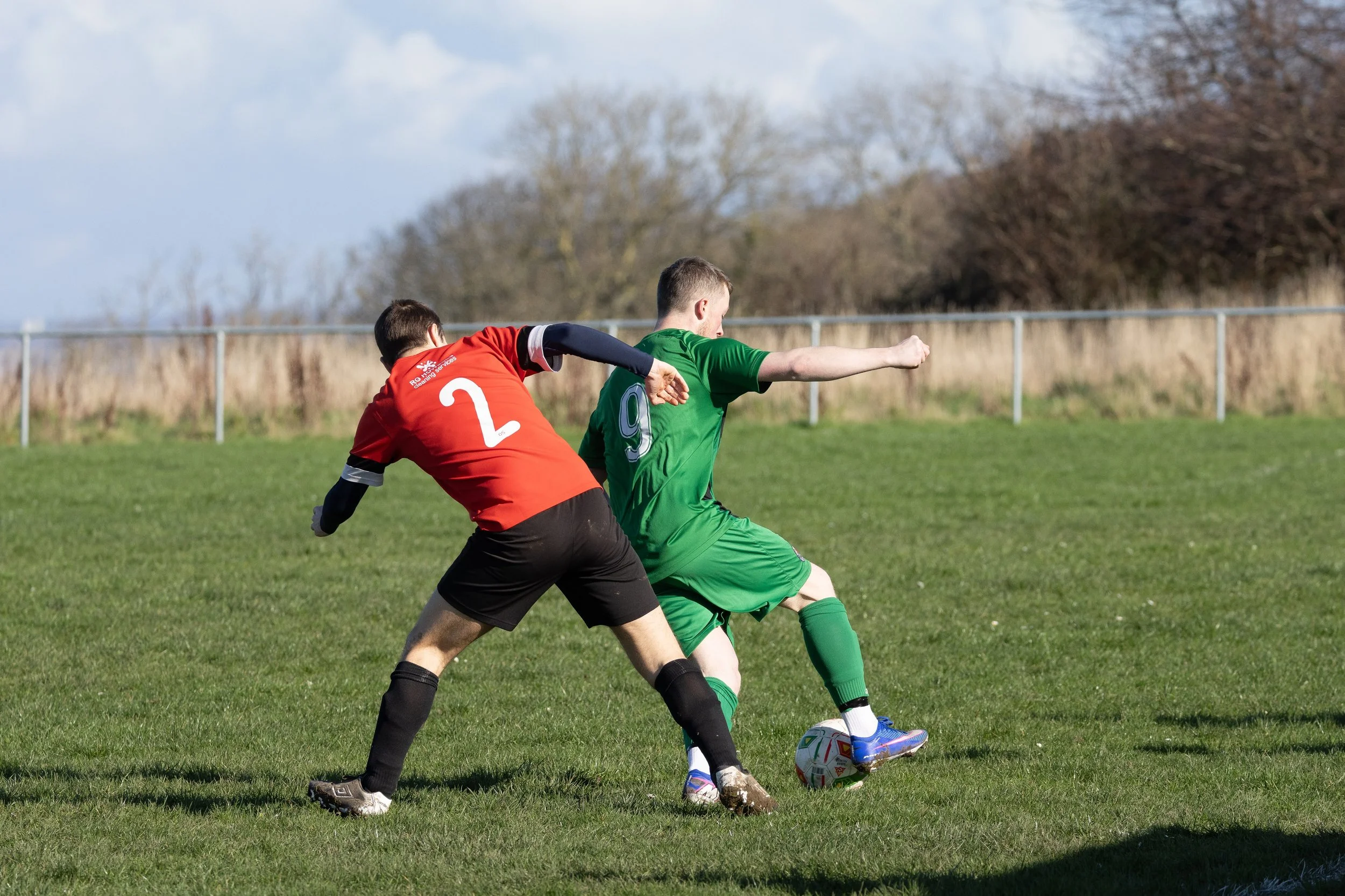 Two soccer players competing for the ball on a grassy field, with a fence and trees in the background.