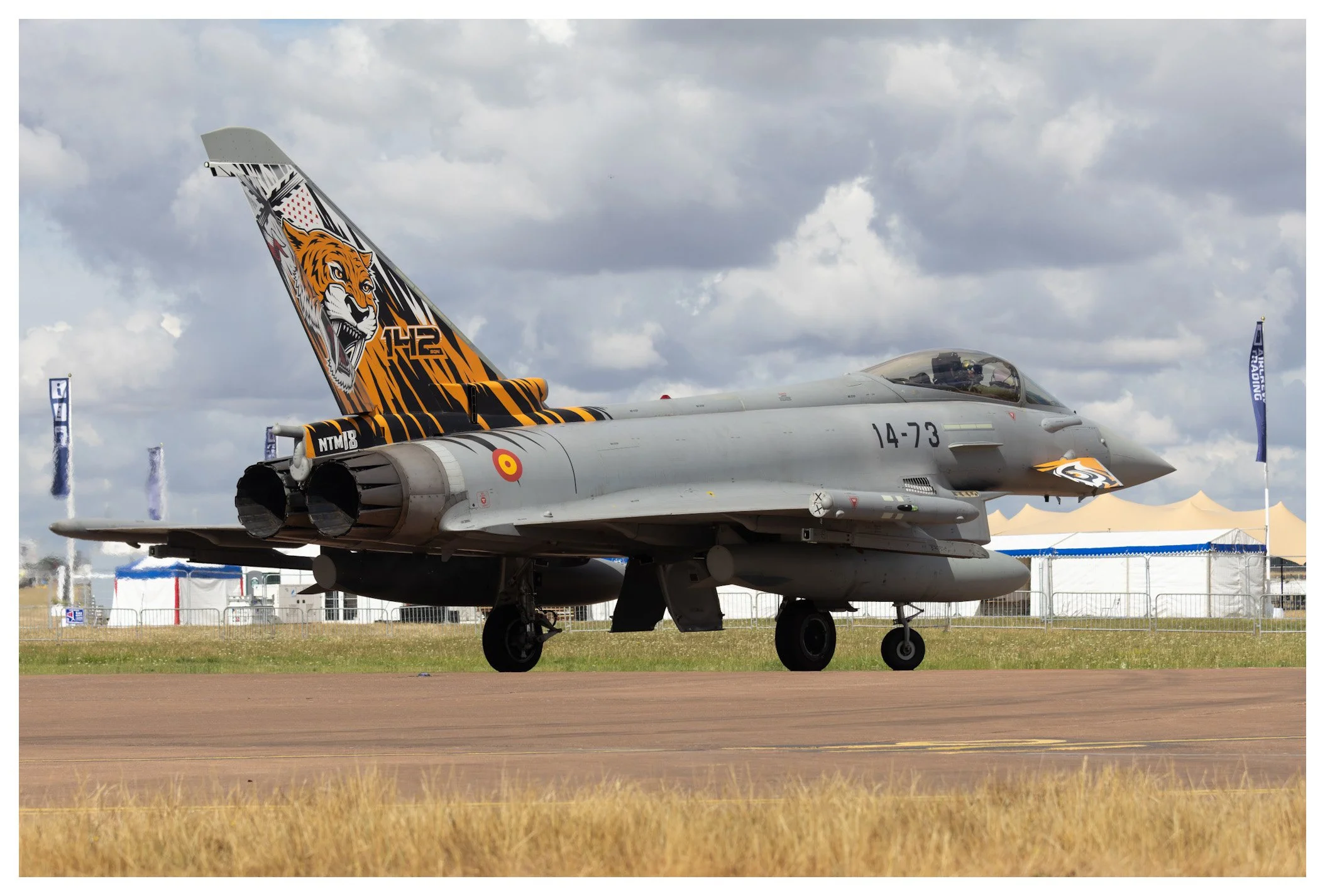 A military fighter jet on the runway with a tiger face design on its tail fin and the number 14-73. The jet is parked on a tarmac at an airshow, with tents and flags in the background.