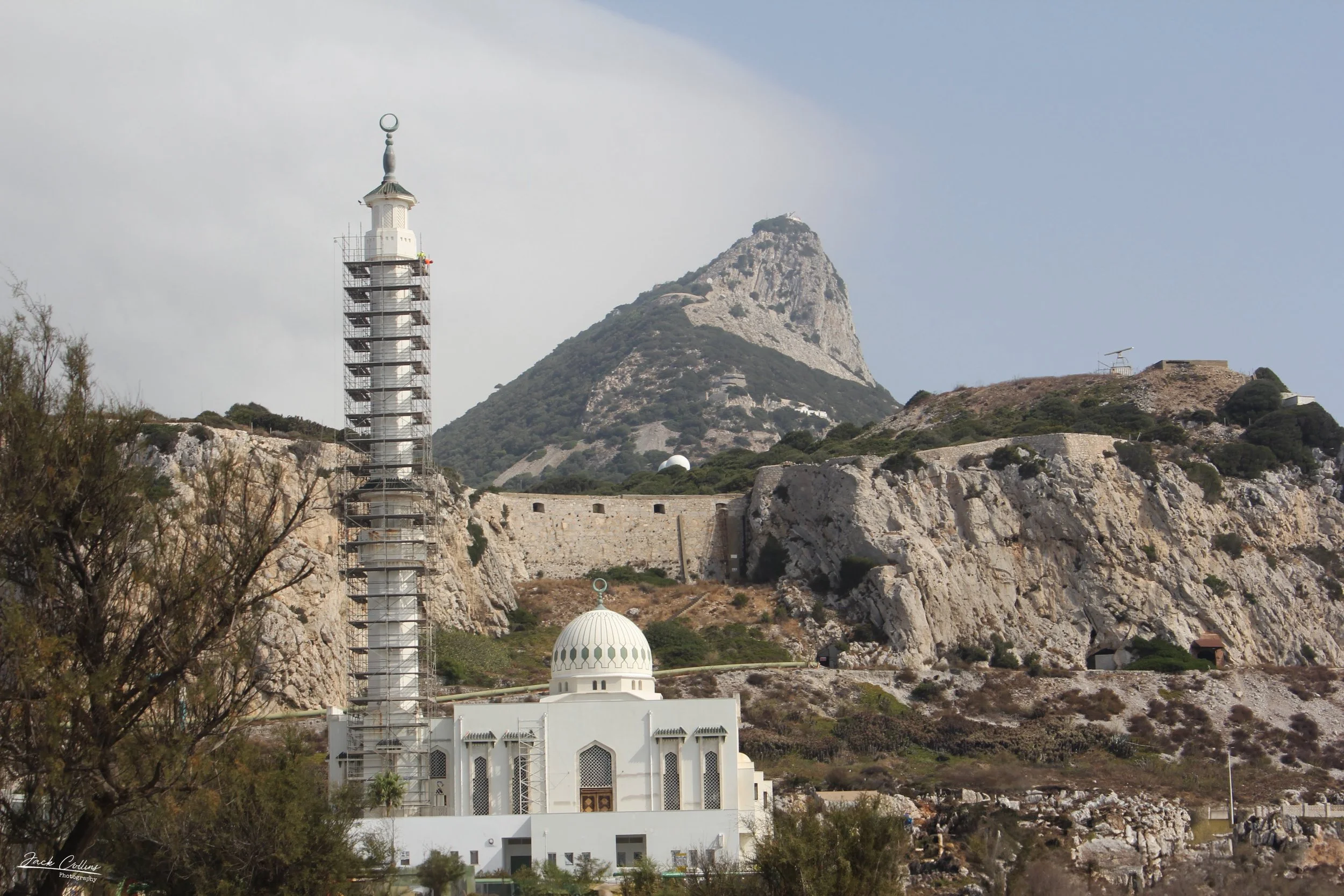 A white mosque with a domed roof and minaret under renovation, with scaffolding around the minaret, set against rocky hills and a mountain in the background.