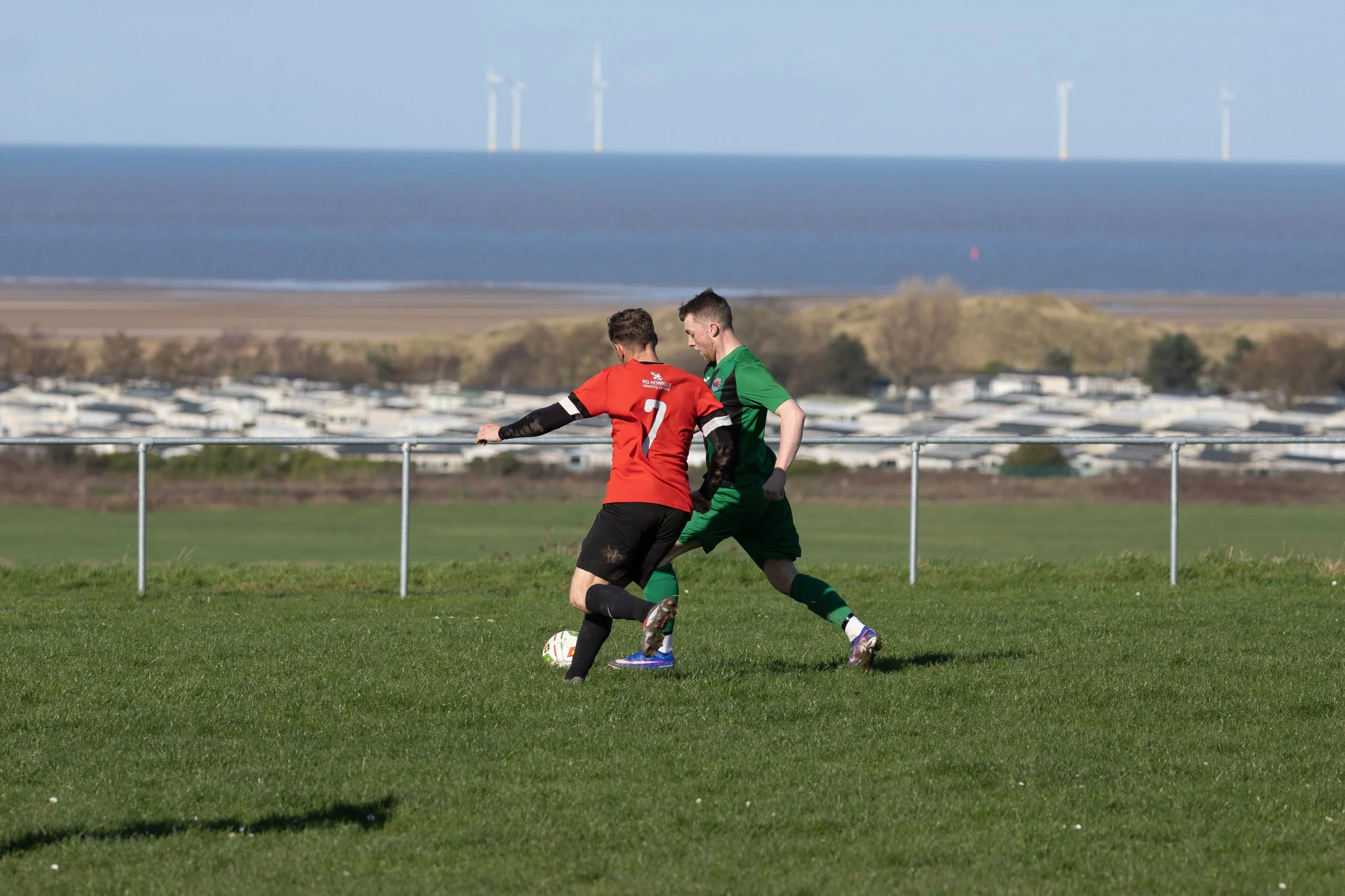 Two soccer players in red and green uniforms compete for the ball on a grassy field with a scenic backdrop of water, land, and wind turbines.
