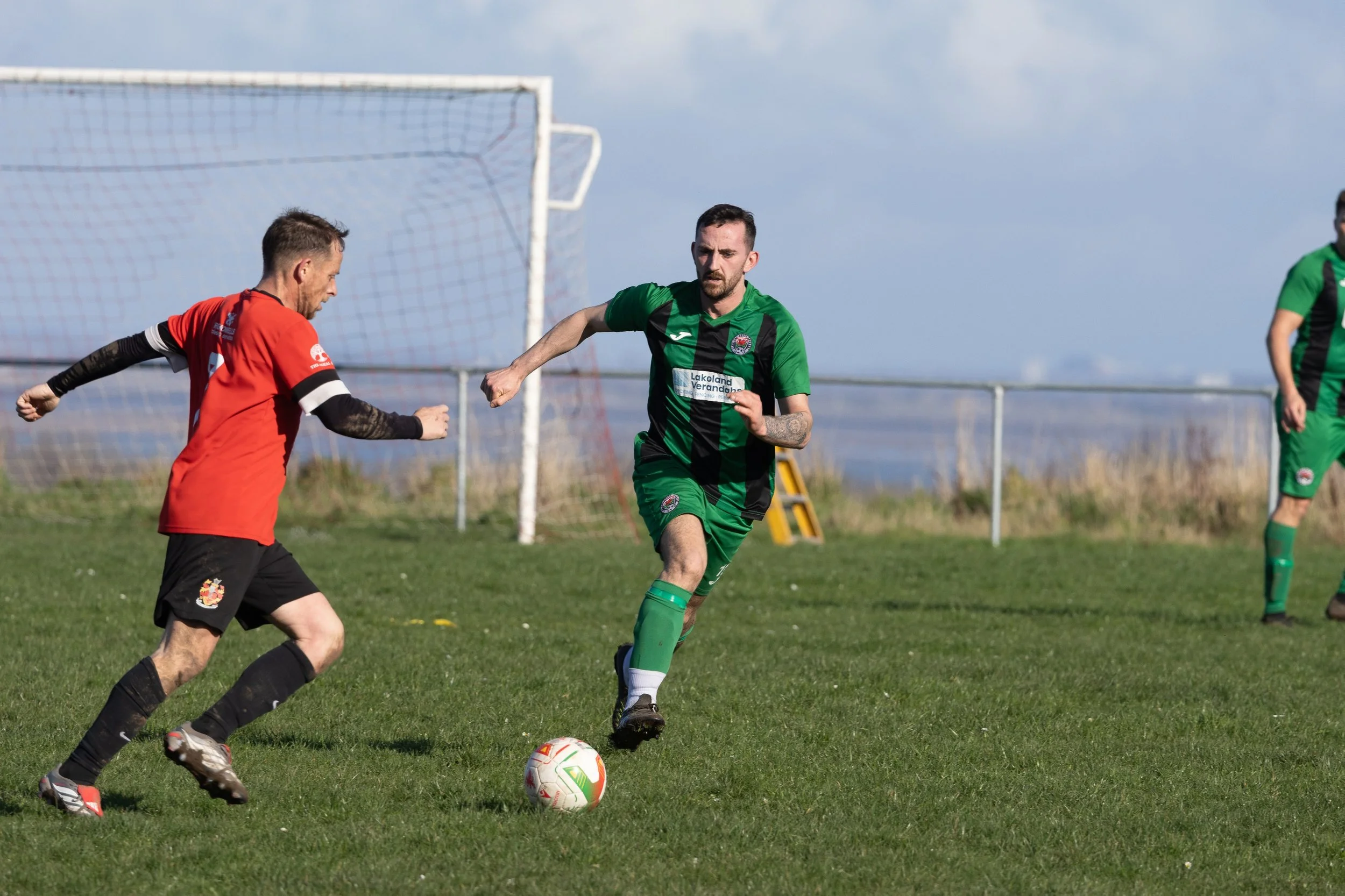 Two soccer players compete for the ball on a grassy field near the goal. One player in a red jersey and black shorts faces off against a player in a green and black jersey and shorts. A third player in green is partially visible to the right.