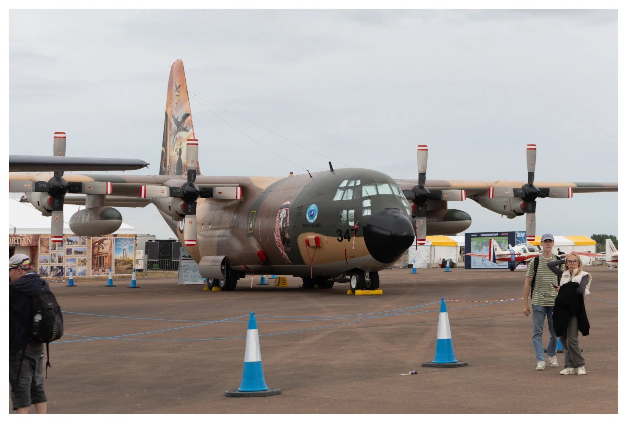 A large military aircraft on display at an airshow, surrounded by blue and white cones with a couple of visitors walking nearby.