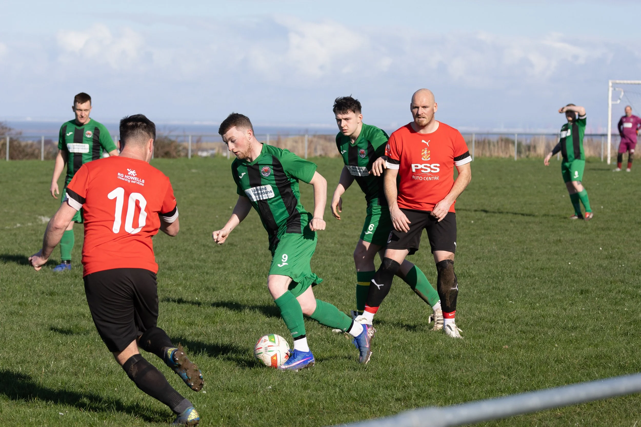 Soccer match with players in green and red jerseys on a grass field, with a goalpost and cloudy sky in the background.