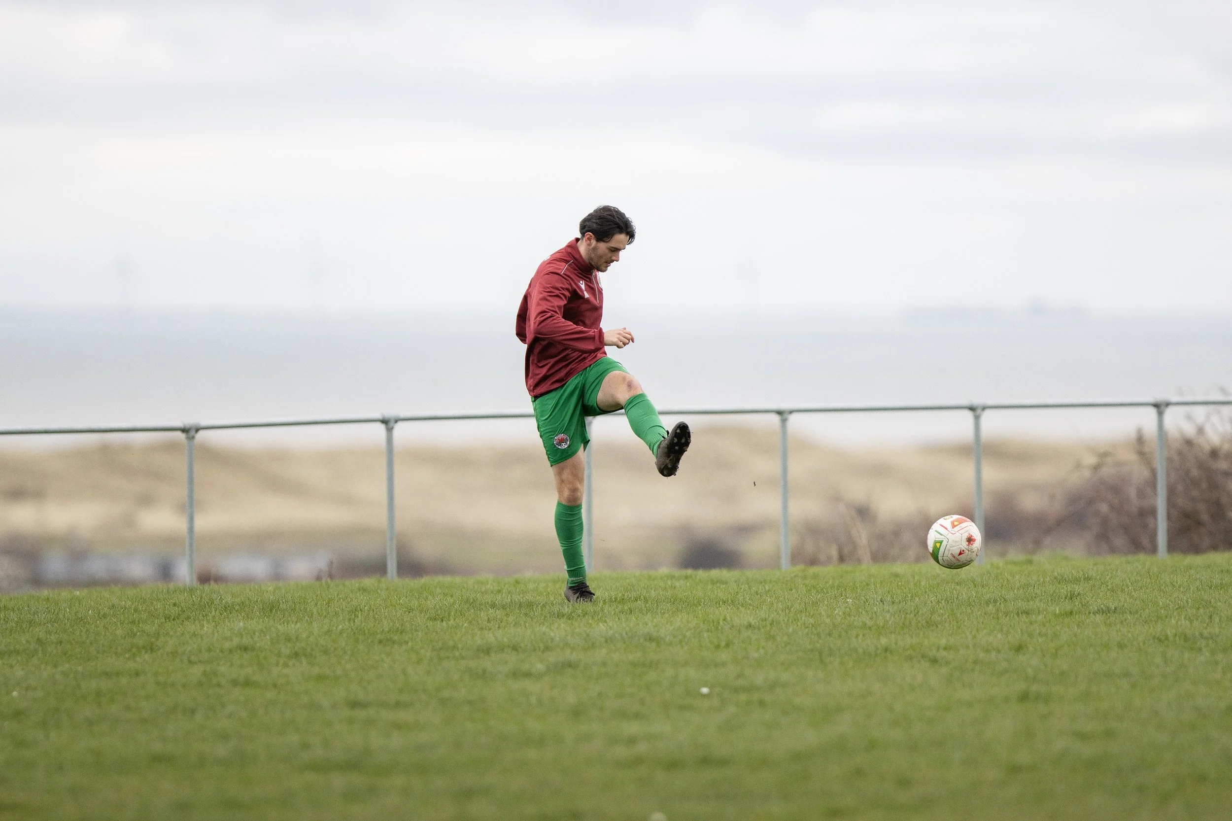 A man in a maroon jacket and green shorts kicking a soccer ball on a grassy field with a metal fence and cloudy sky in the background.