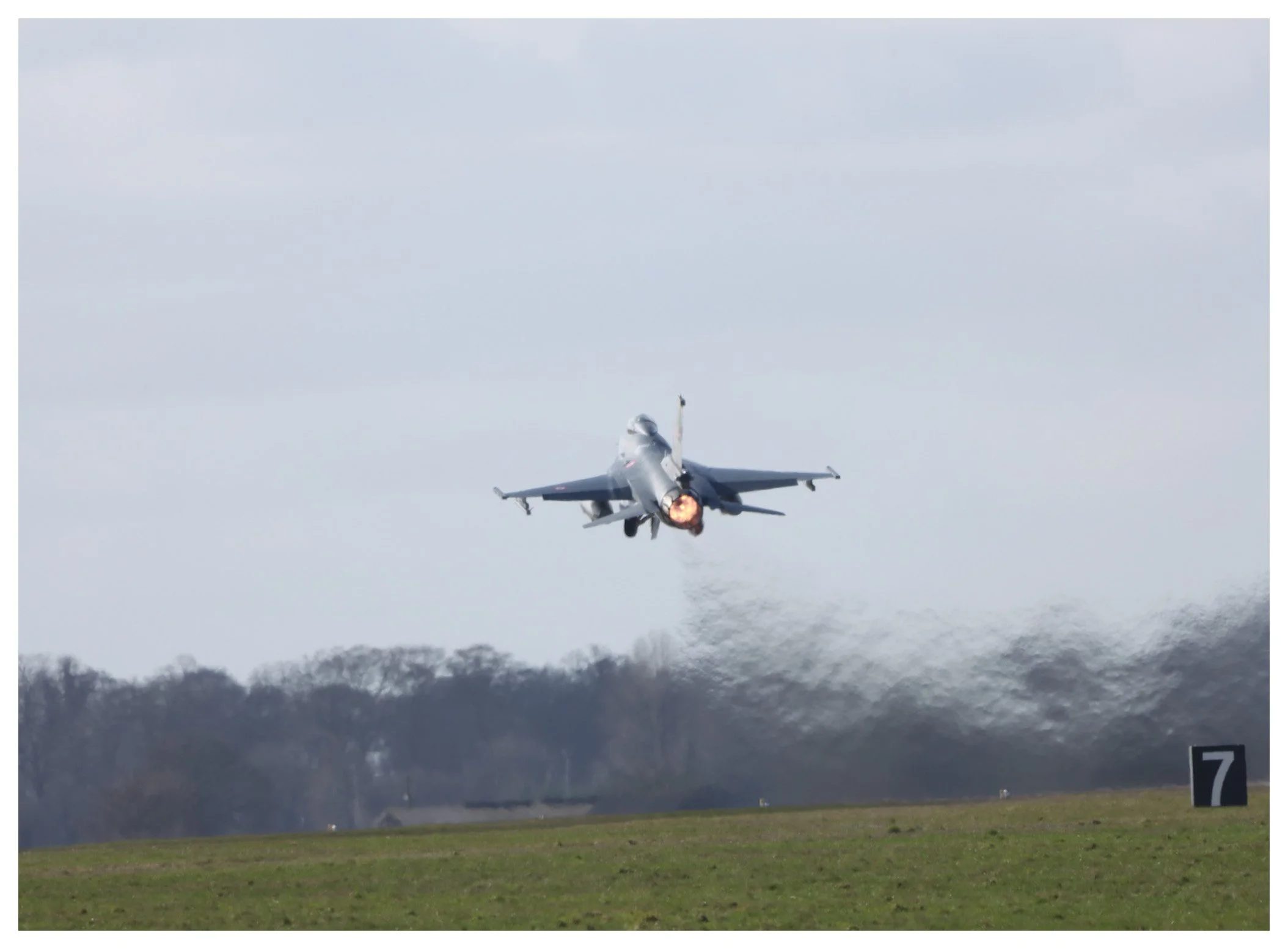 Military jet launching afterburner flying low over a grassy field with a treeline in the background, near a numbered runway marker.