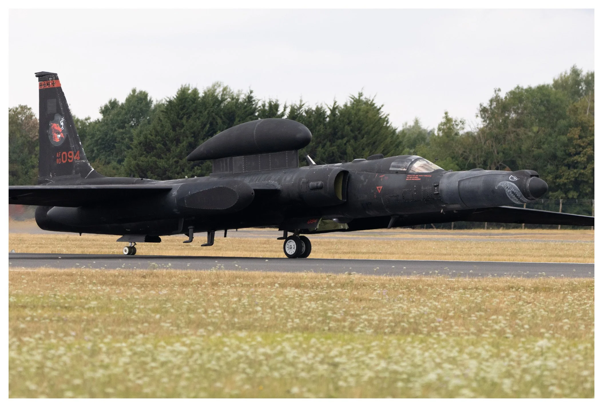 A black military fighter jet airplane on the runway with trees in the background.