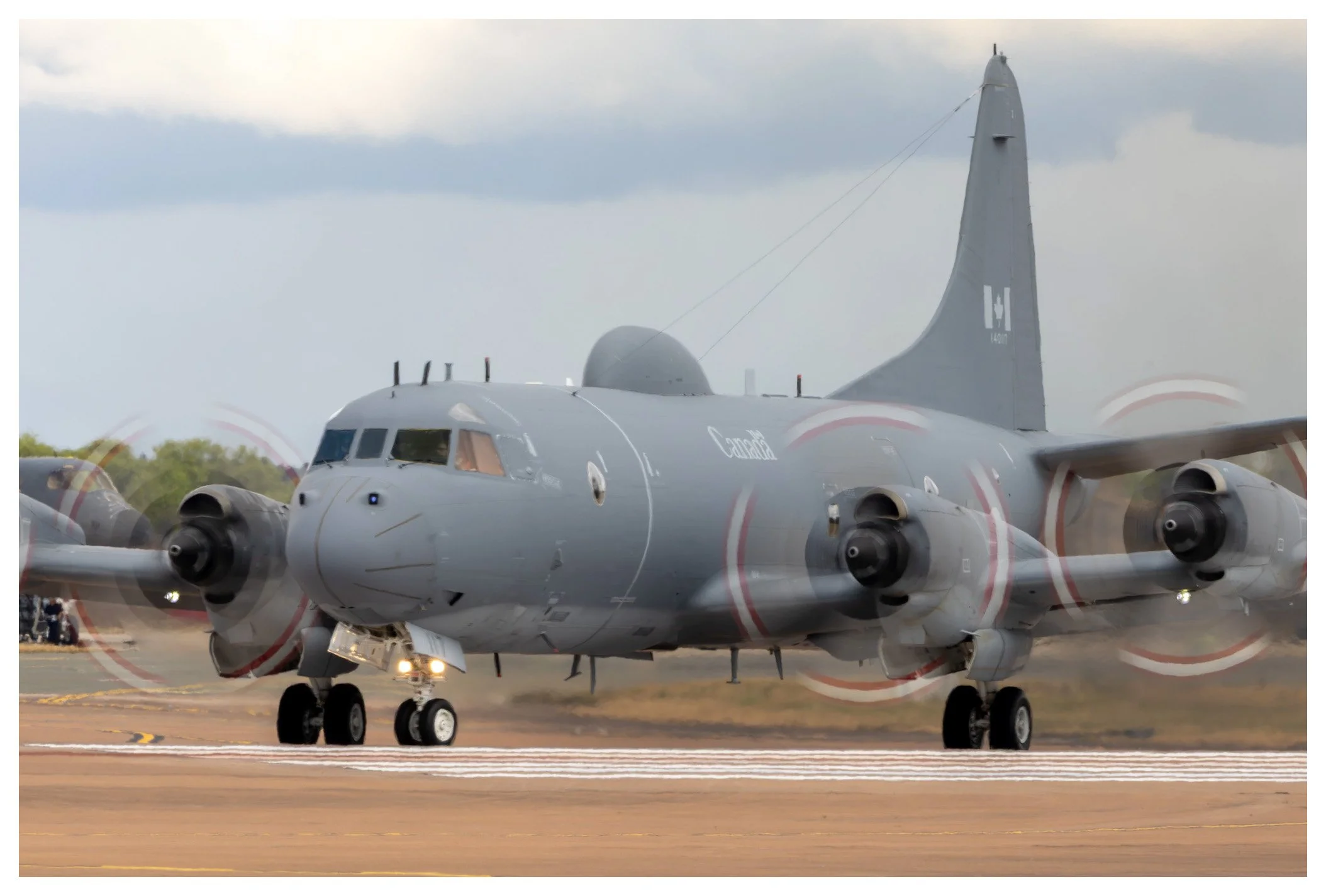 A grey military cargo aircraft with four propeller engines taxiing on a runway with a cloudy sky in the background.