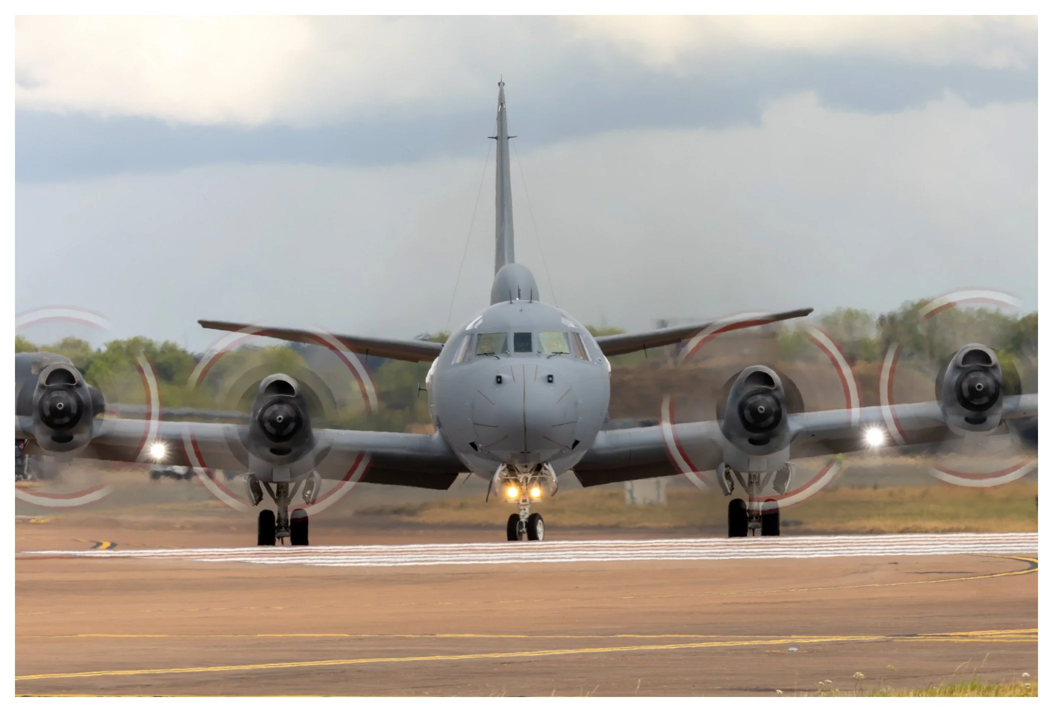 A gray military airplane on the runway with four spinning propellers, preparing for takeoff, with clouds in the sky and greenery in the background.