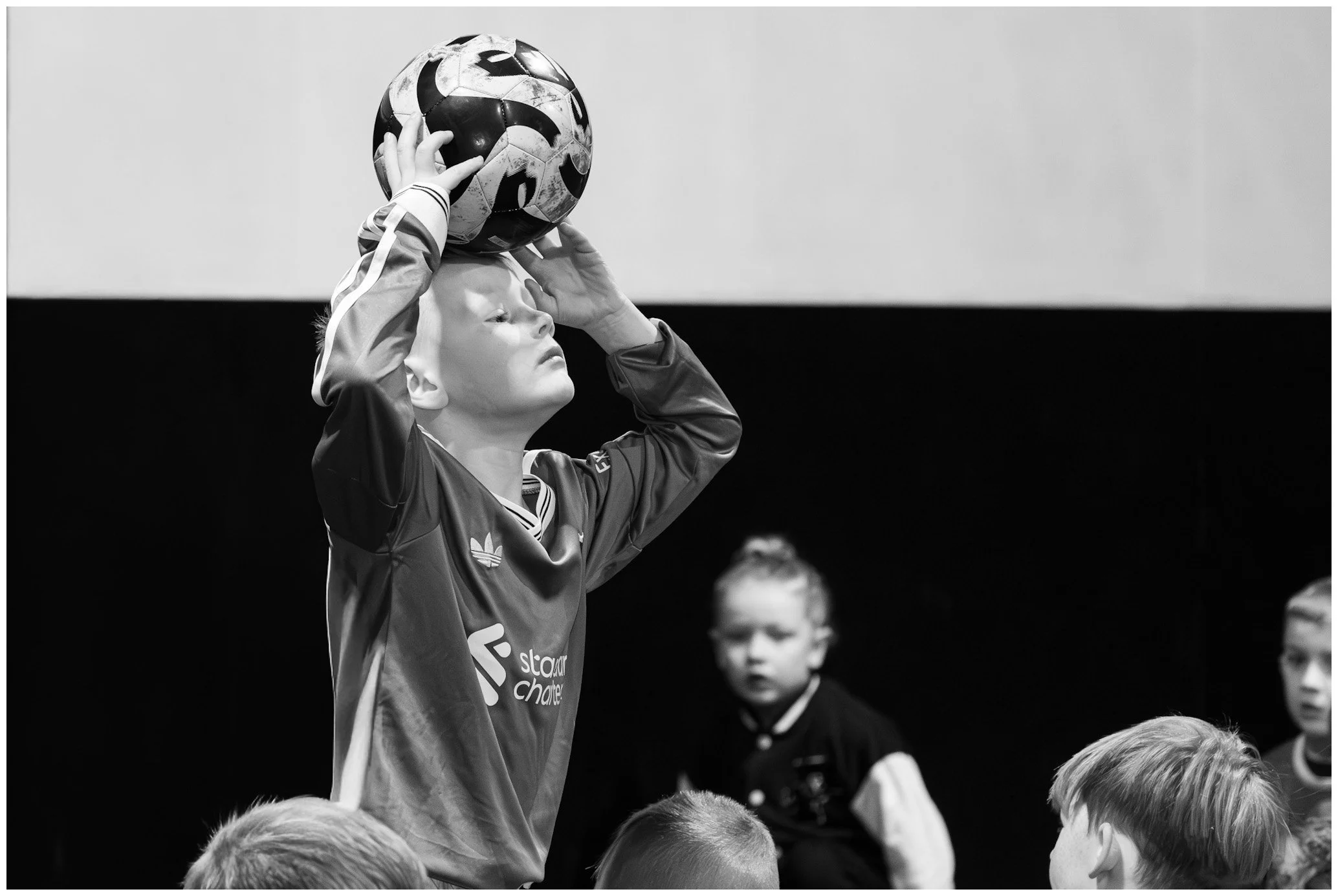 A young girl with blonde hair in a bun holding a soccer ball above her head, preparing to throw or catch it, while several boys watch nearby.
