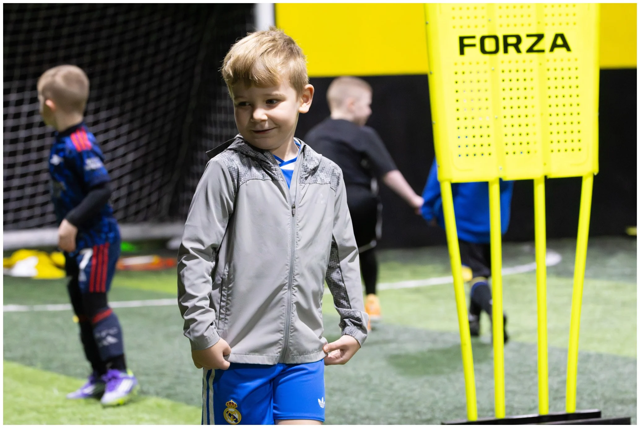 Young boy in a gray jacket and blue shorts standing on an indoor soccer field, with two other children in the background practicing near a yellow training obstacle labeled 'FORZA'.