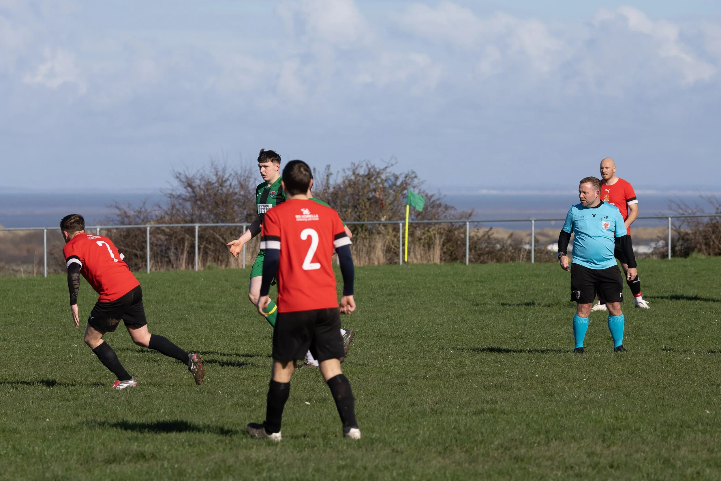 Soccer match with players in red and green jerseys on a grassy field, and a referee in a light blue shirt, during daytime with partly cloudy sky.