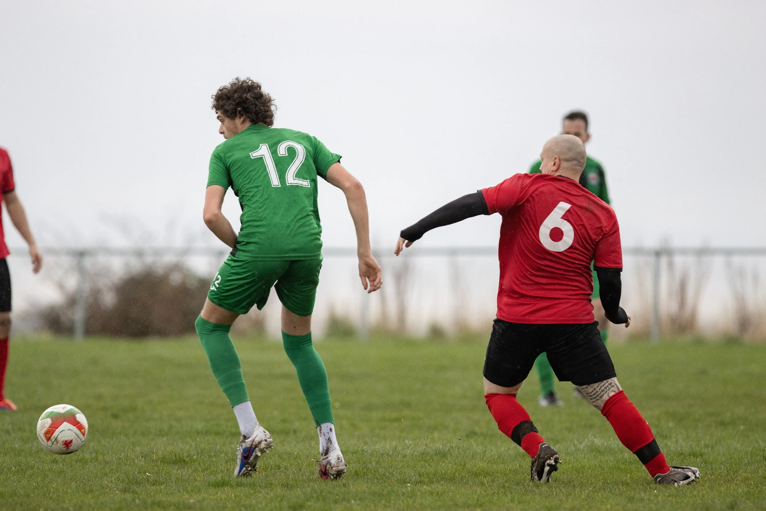 Soccer players competing for ball, one in green jersey with number 12, another in red jersey with number 6, on a grassy field with overcast sky.