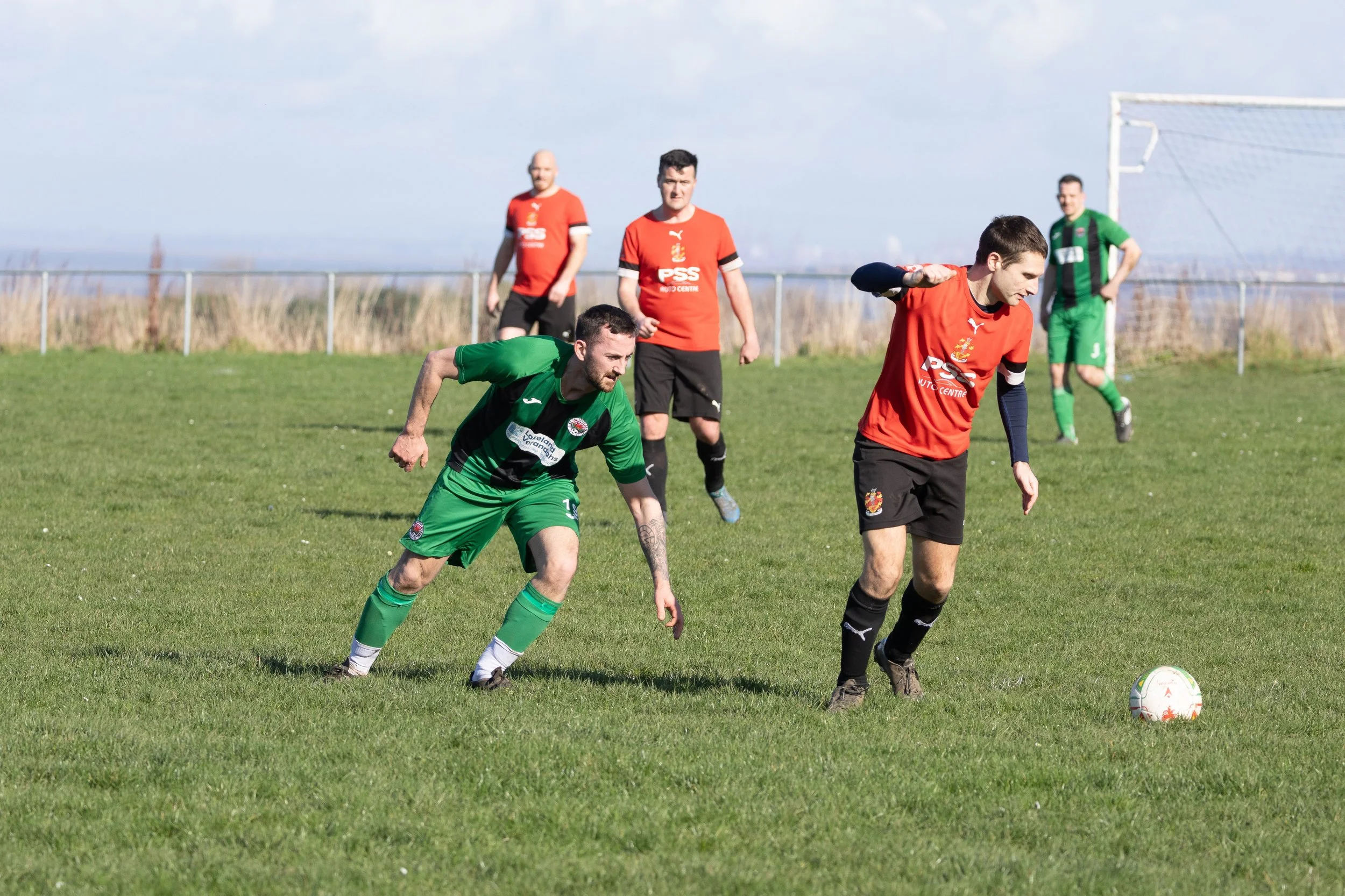 Four soccer players on a green field, with one in red preparing to kick a ball, one in green rushing towards the ball, and two others in red and green standing in the background near the goal. Clear sky, rural landscape.