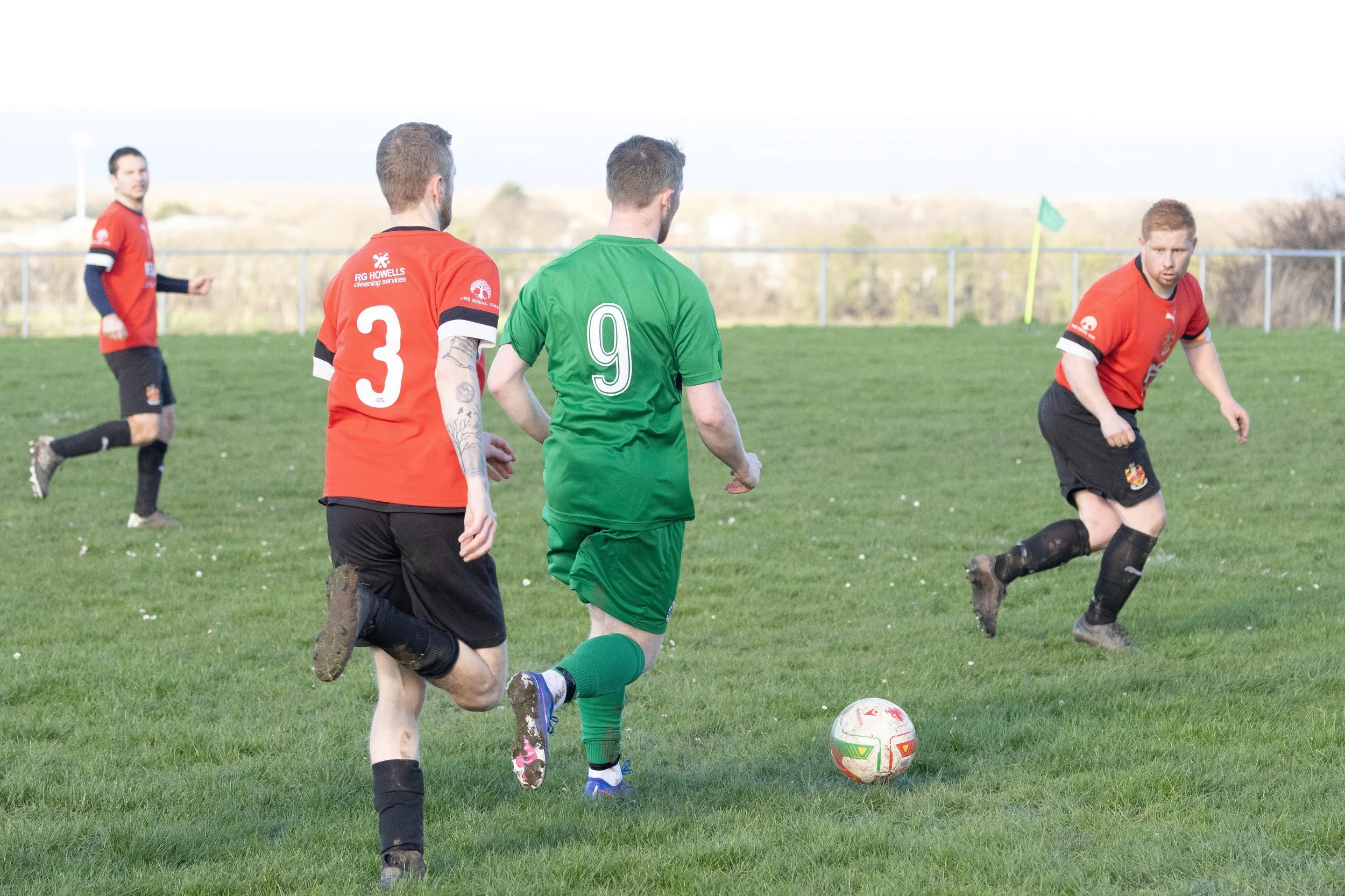 Group of soccer players on a green field during a match, with one player in a green uniform and others in red and black uniforms, some running towards a soccer ball.