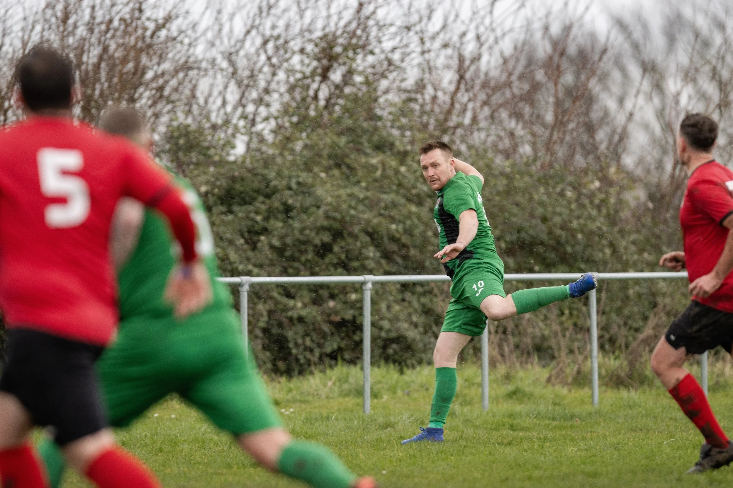 Soccer match with players in green and red jerseys on grass field, focusing on a player in green preparing to kick the ball, with other players blurred in the foreground and background, and trees in the distance.