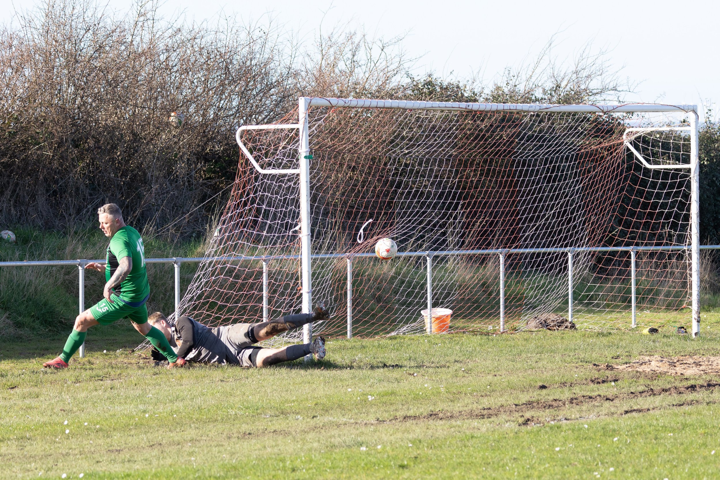 A soccer game scene with a player in a green jersey walking past a fallen goalie in gray and black gear on the grass, a ball in mid-air near the net, and a goalpost with orange netting in the background.