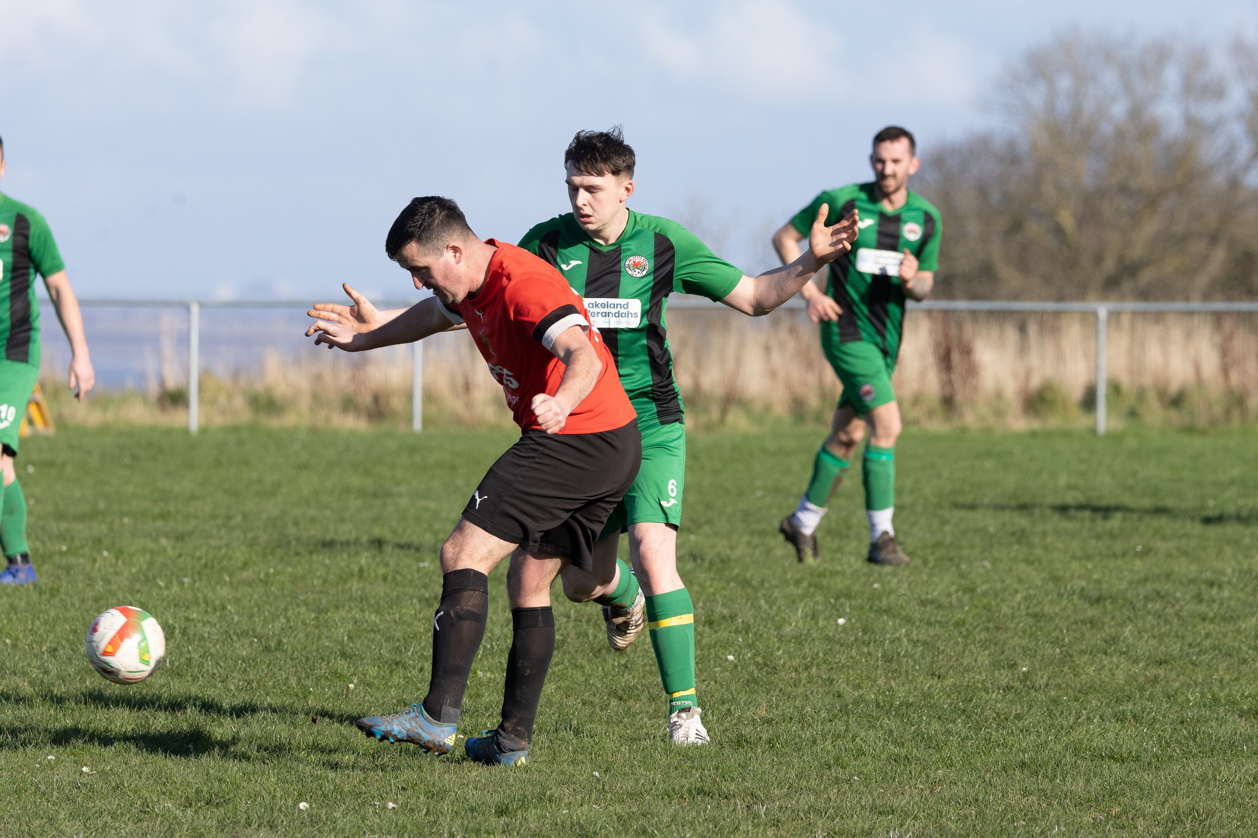 Soccer players competing for the ball on a grassy field with a metal fence and trees in the background.