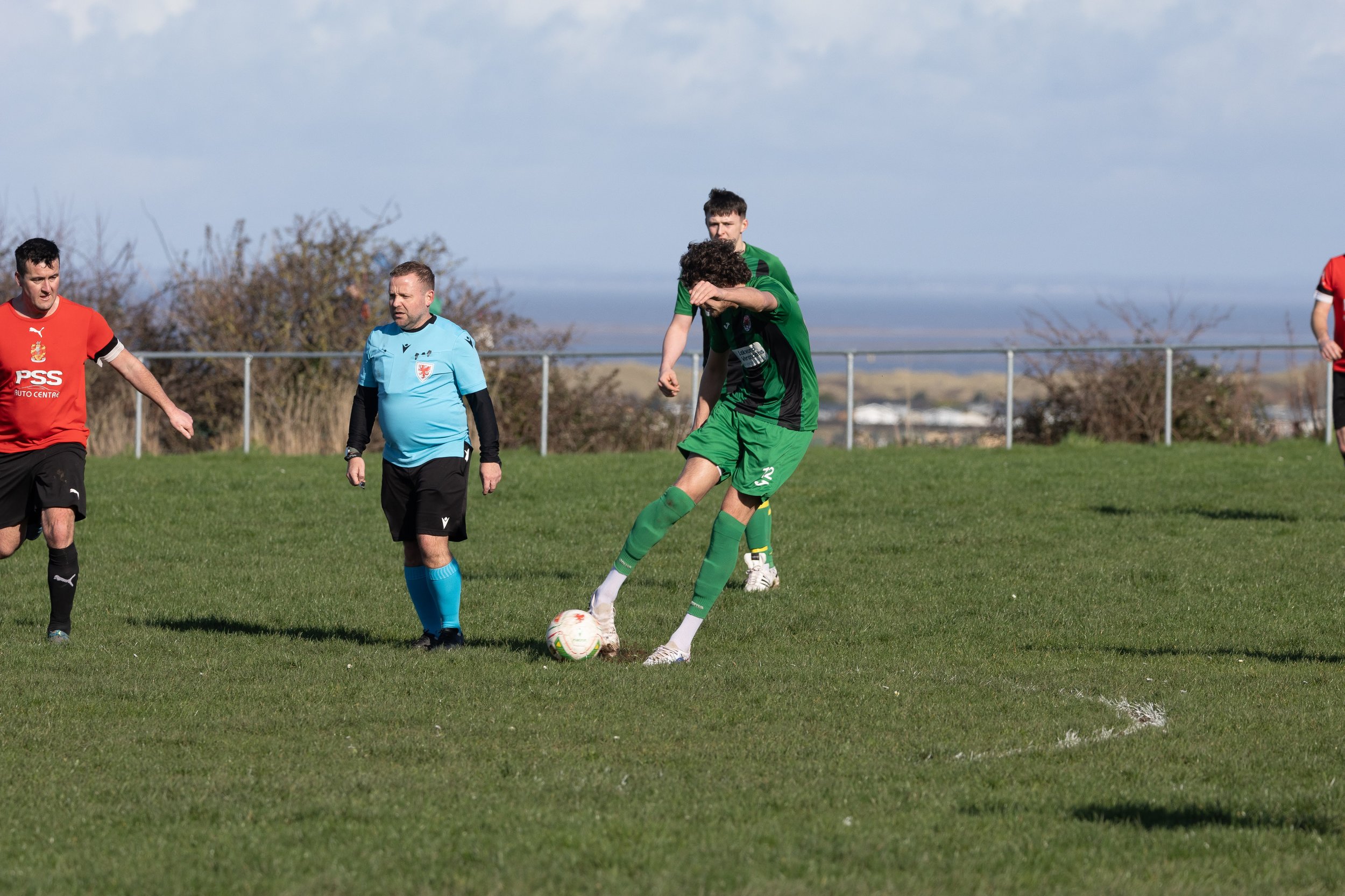 Soccer match with players and referee on a grassy field under a blue sky.