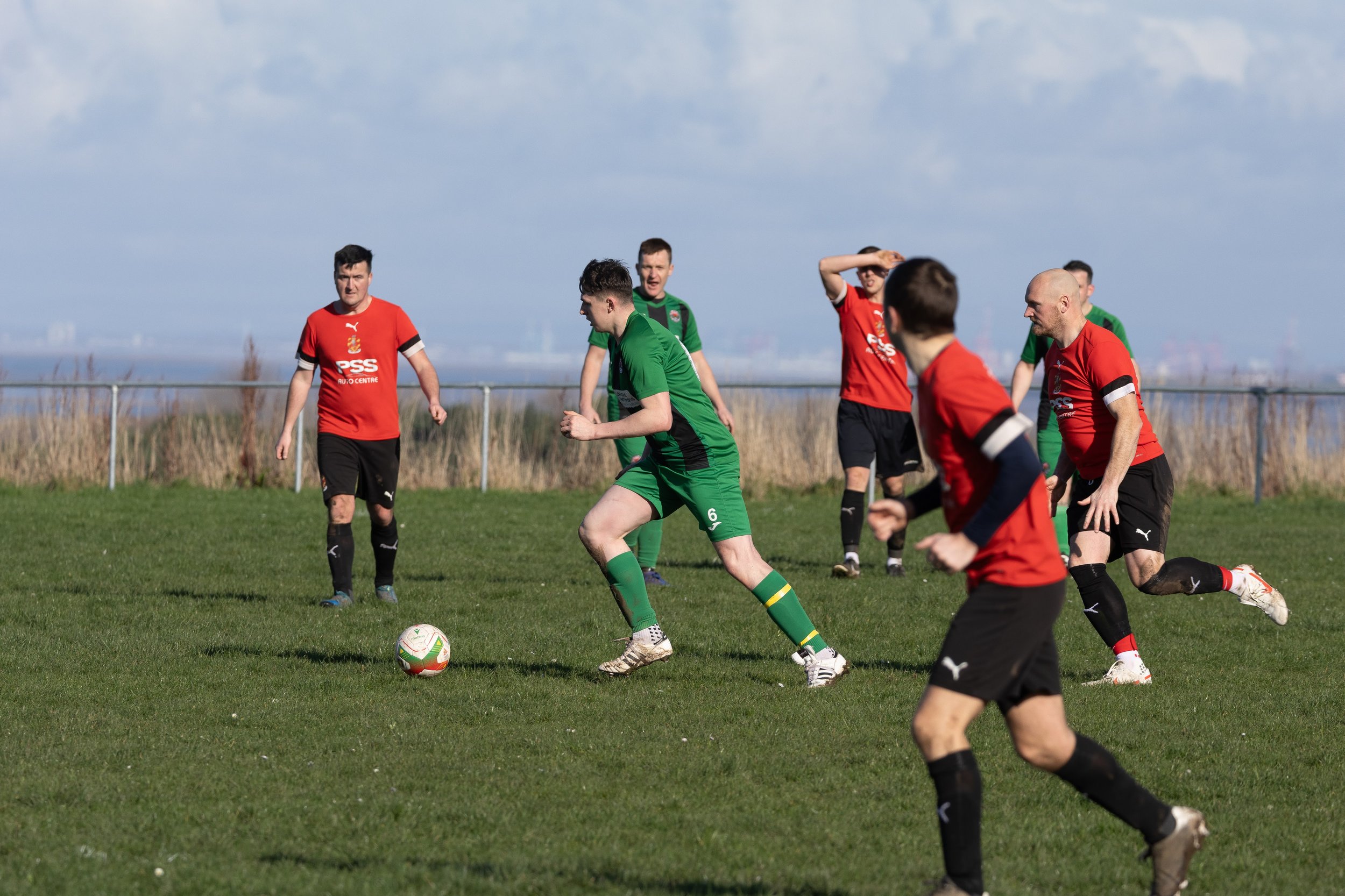 Soccer players on a field during a match, wearing red and green jerseys, with a ball on the grass and a blue sky with clouds in the background.
