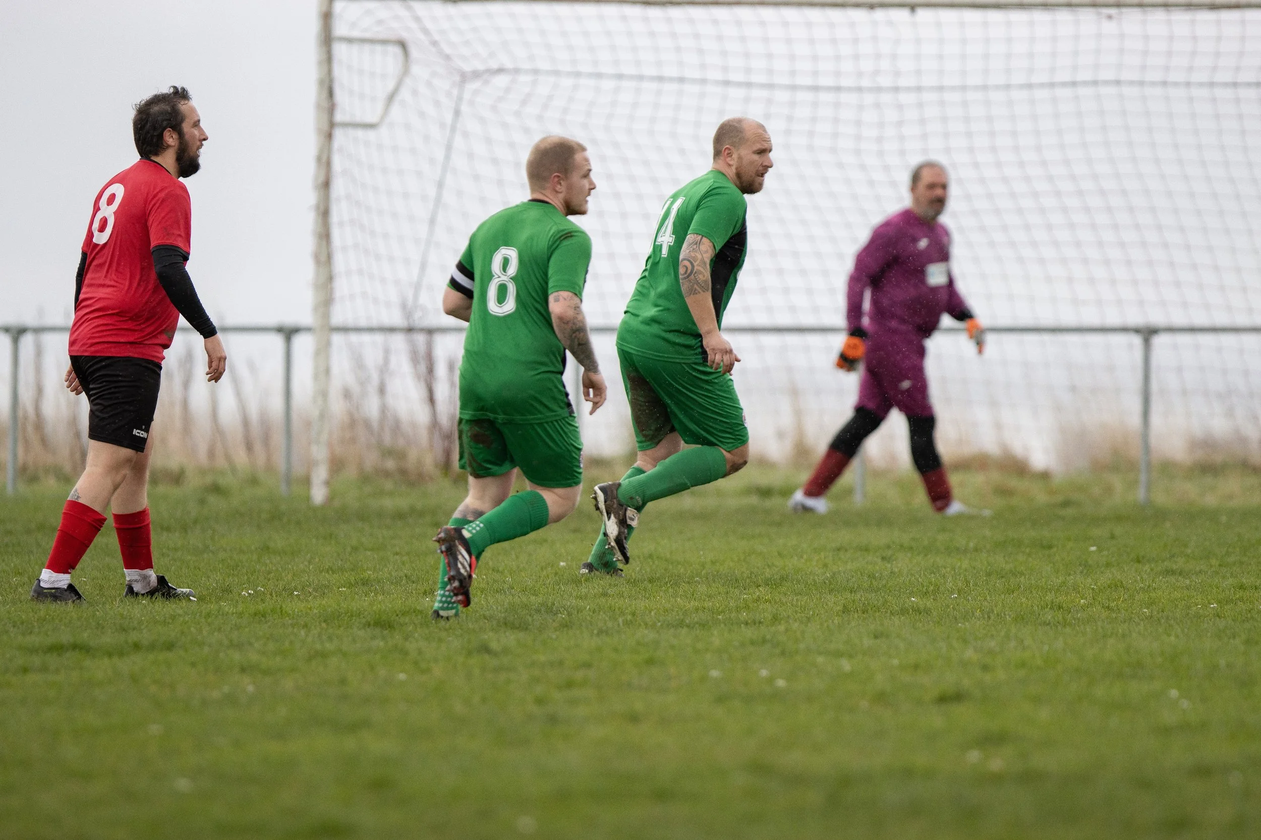 Soccer players on the field during a game, with two players in green jerseys, one in a red jersey, and a goalkeeper in a purple uniform