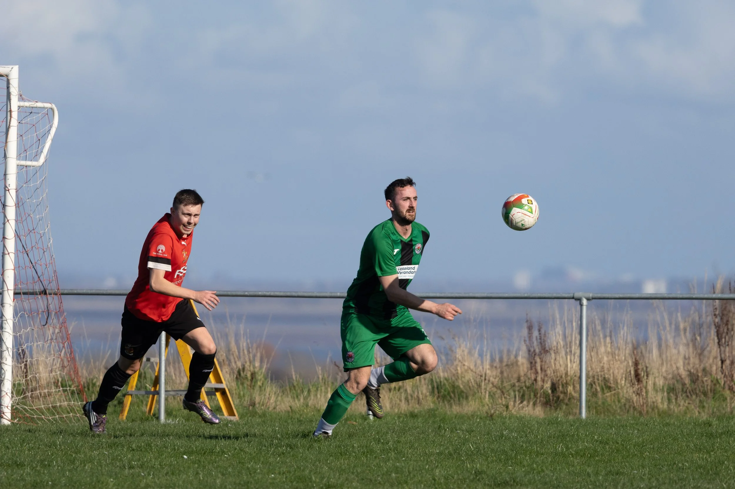 Two soccer players in action on a grassy field with a goal and mesh net, one in a red jersey and the other in a green jersey, during a daytime match under a partly cloudy sky.
