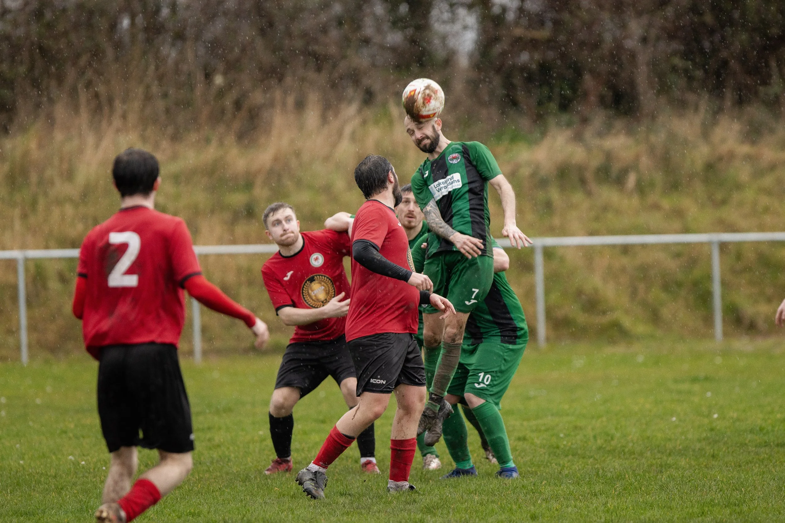 Soccer players in red and green uniforms contest an aerial ball during a match on a rainy day.
