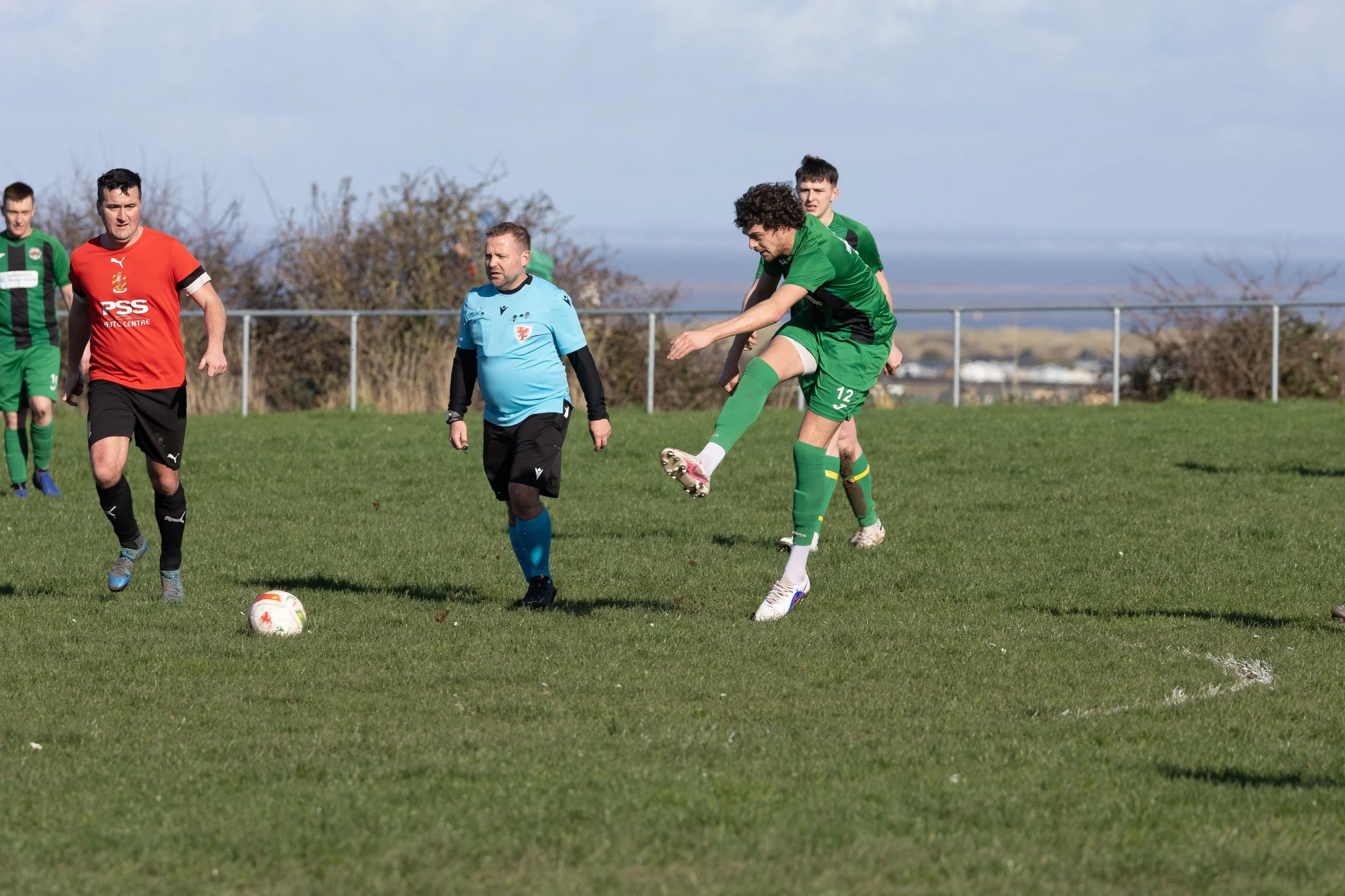 A soccer match with players in green, red, and black uniforms on a grassy field, with a referee in a blue shirt, during daytime with clear weather.