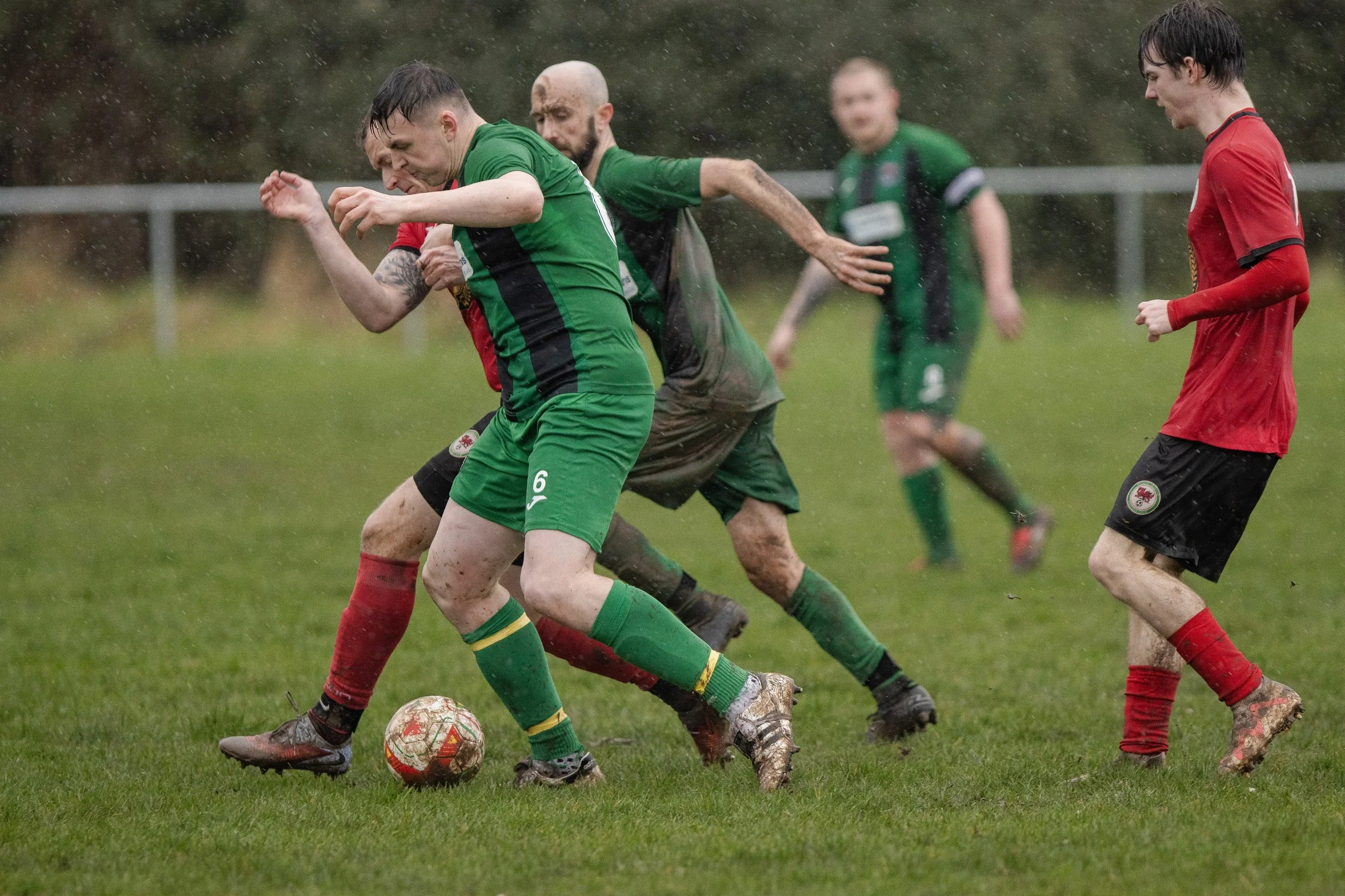 A soccer match in rain with players in green and red jerseys competing for the ball on grass field.
