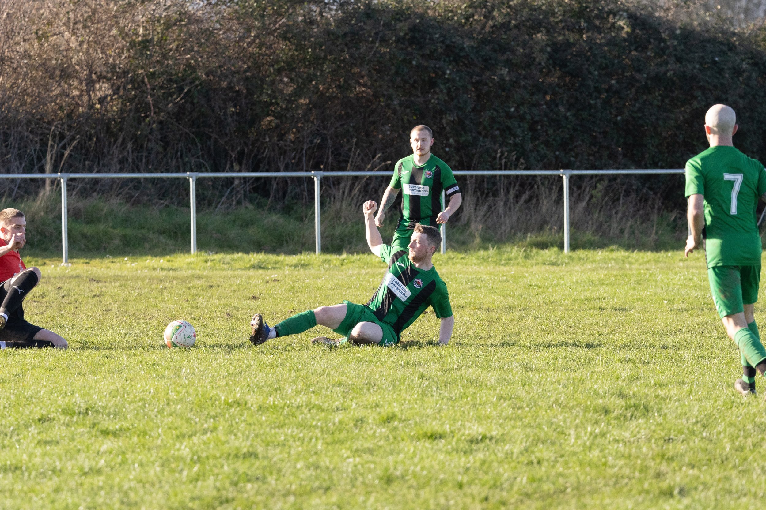 Soccer players during a game on a grassy field, with one player sitting on the ground and celebrating while others are nearby.