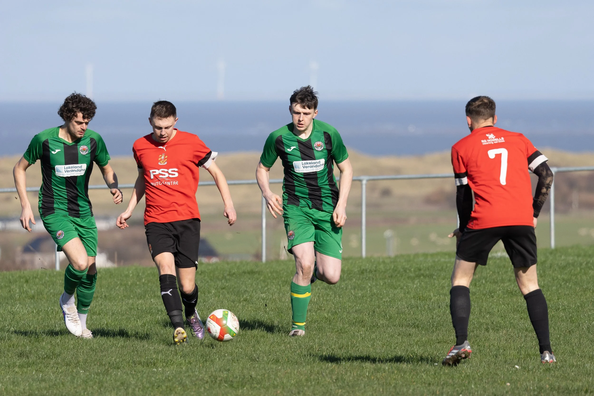 Four young male soccer players on a grassy field during a game. Two players are in green jerseys and two in red, with one in the middle controlling the ball. The background shows a landscape with hills and a body of water under a clear sky.