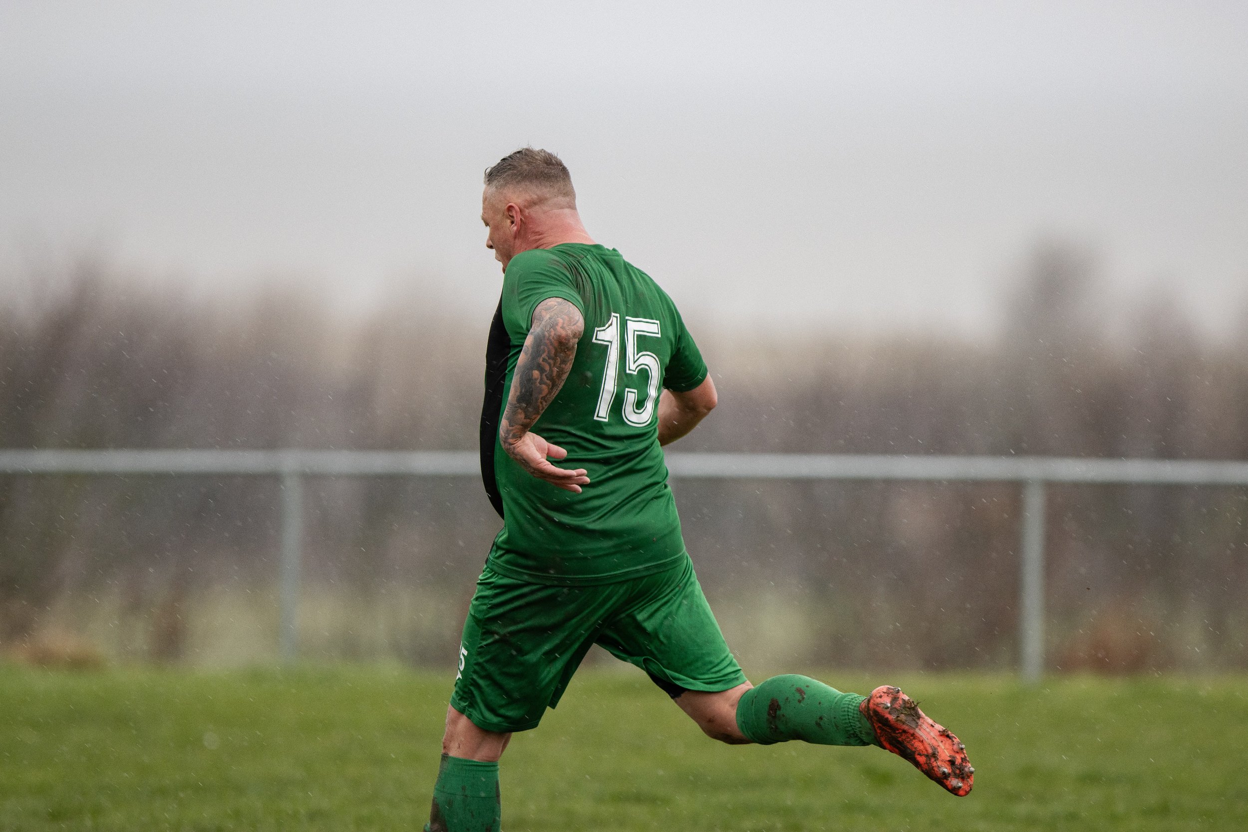 Soccer player wearing a green uniform with the number 15, kicking a ball on a grassy field in rainy weather.