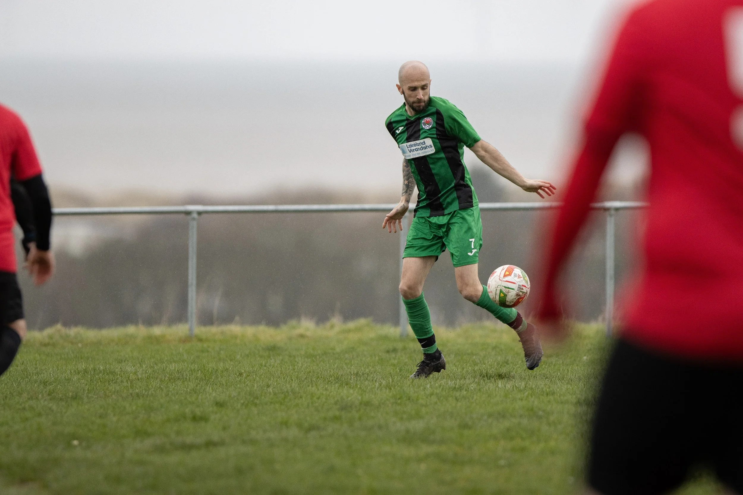 A soccer player with a bald head, beard, and tattoos is wearing a green and black uniform, kicking a white soccer ball on a grassy field during a match.