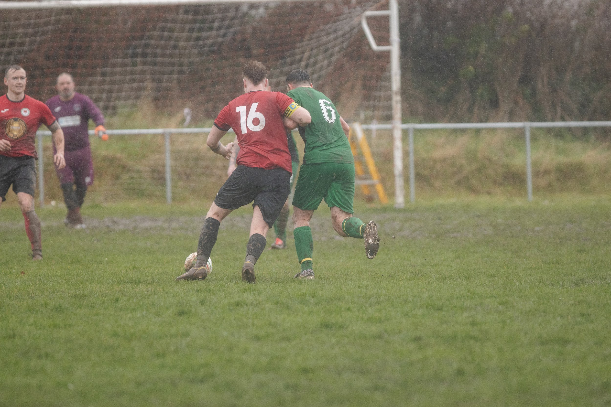 Soccer players in muddy field during match, two players contesting for ball, others watching. Overcast weather, wet grass.